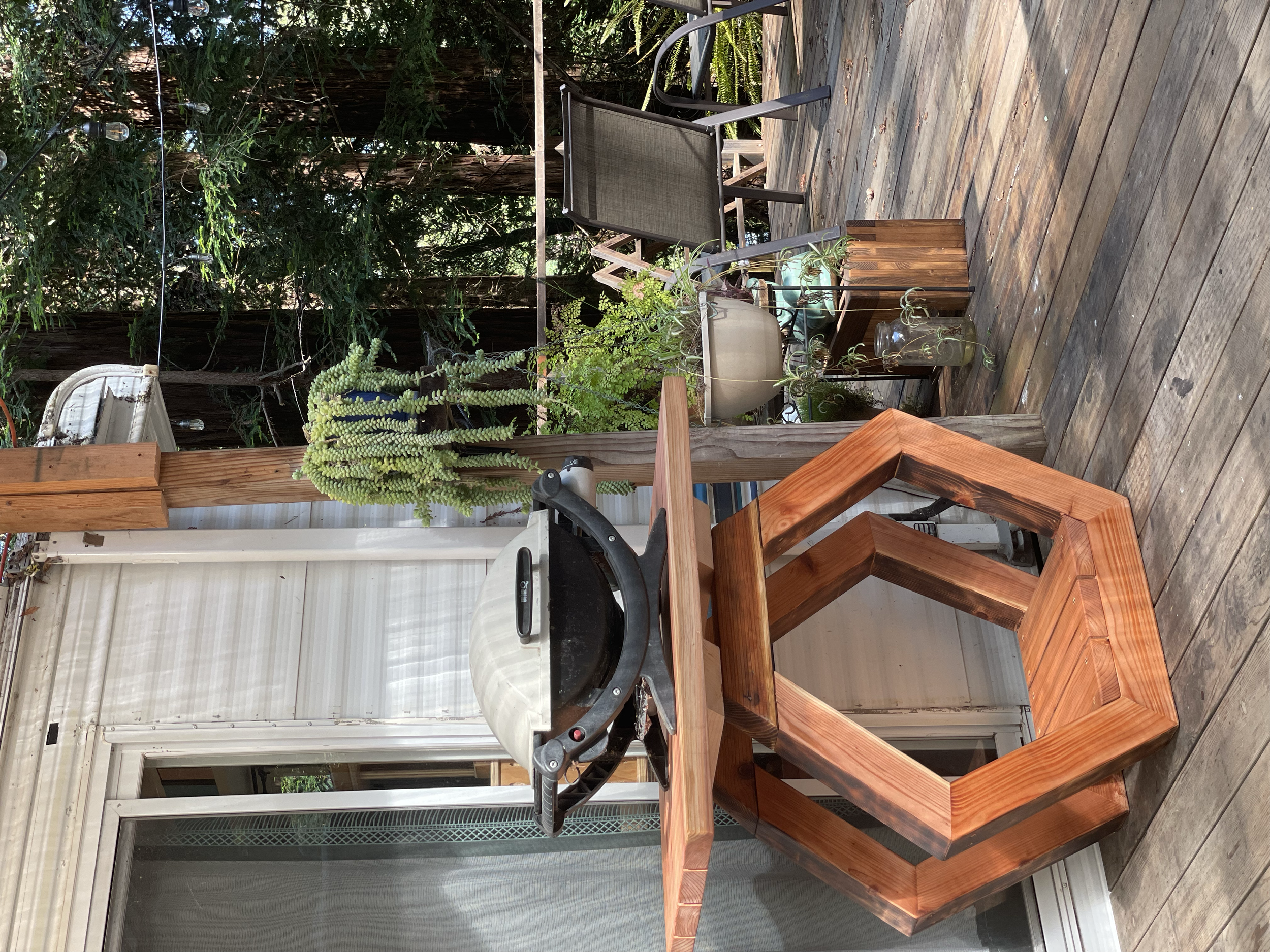 Outdoor wooden deck with a small round table, a gray lounge chair, and potted plants. A grill is on the table, and a window with a white frame is on the house in the background. There are tall trees and greenery surrounding the patio.