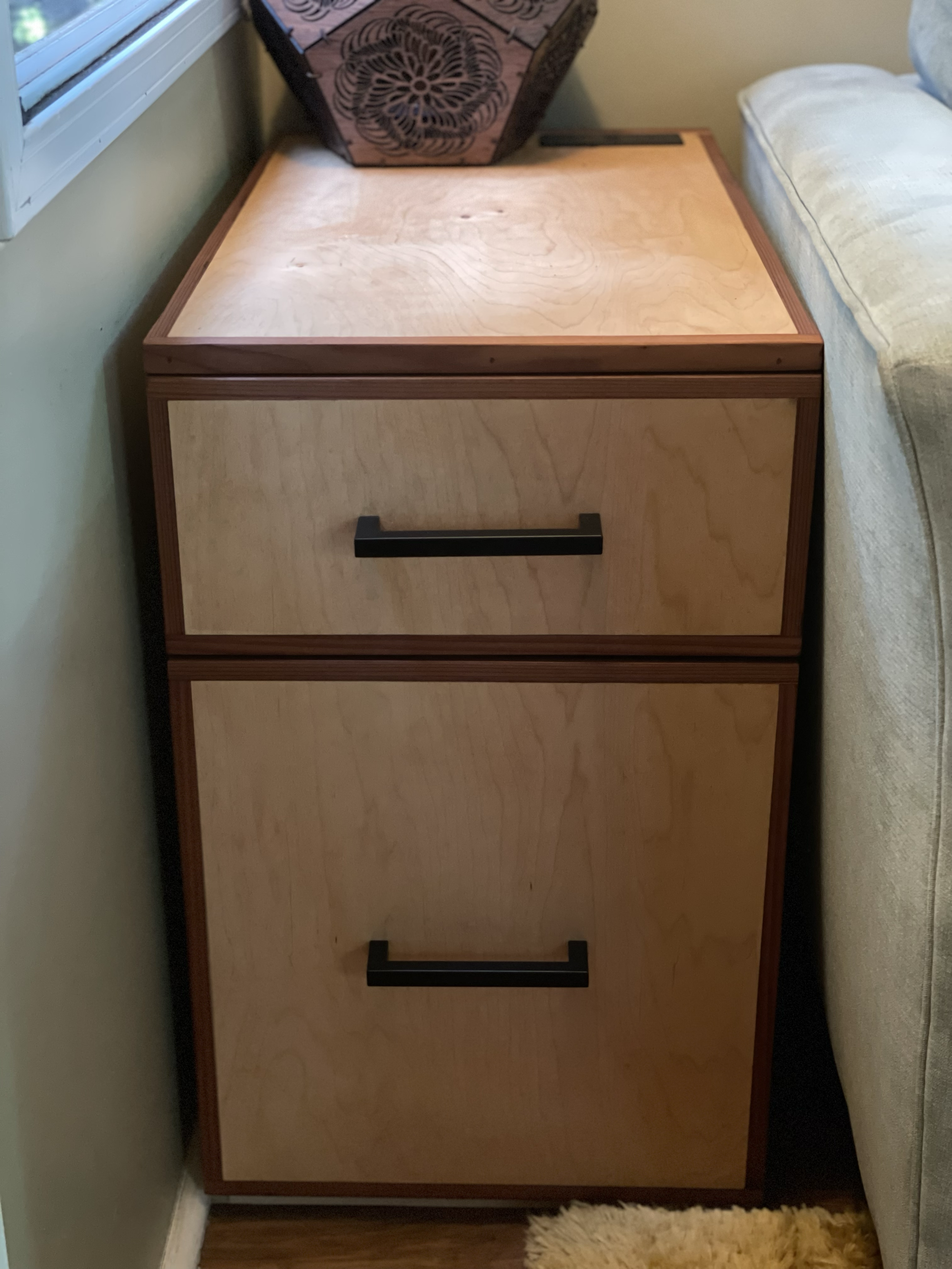 Two-tier wooden filing cabinet with black handles, positioned beside a beige couch and a window with white trim.