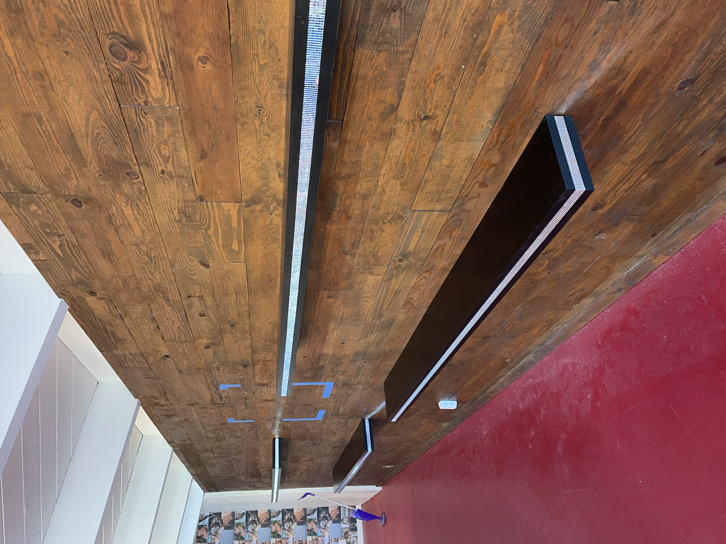 Wooden ceiling with built-in lighting fixtures and electrical outlets, in a room with red painted floor and partially visible white wall with steps.