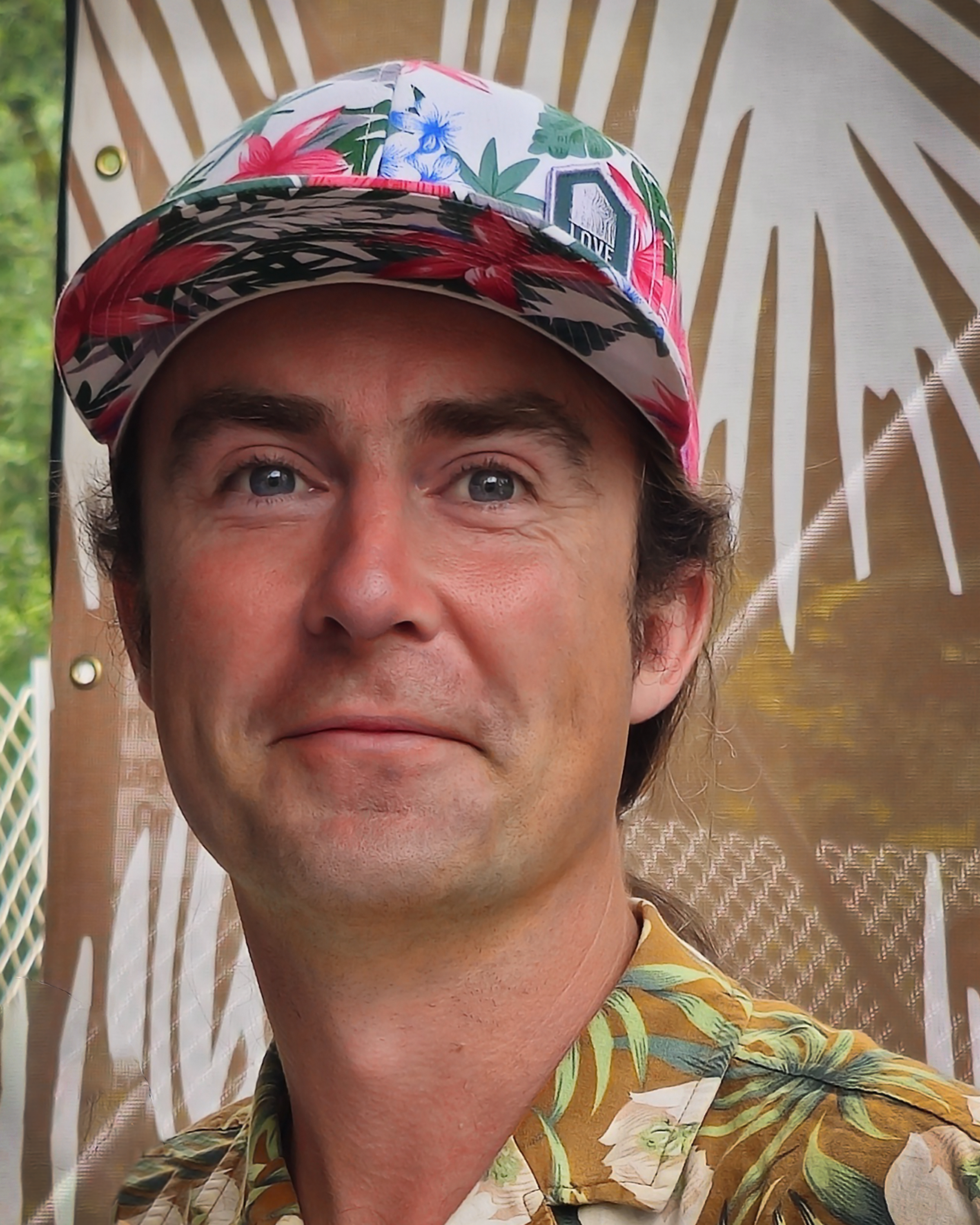 A man with blue eyes and brown hair wearing a colorful floral hat and a Hawaiian shirt.