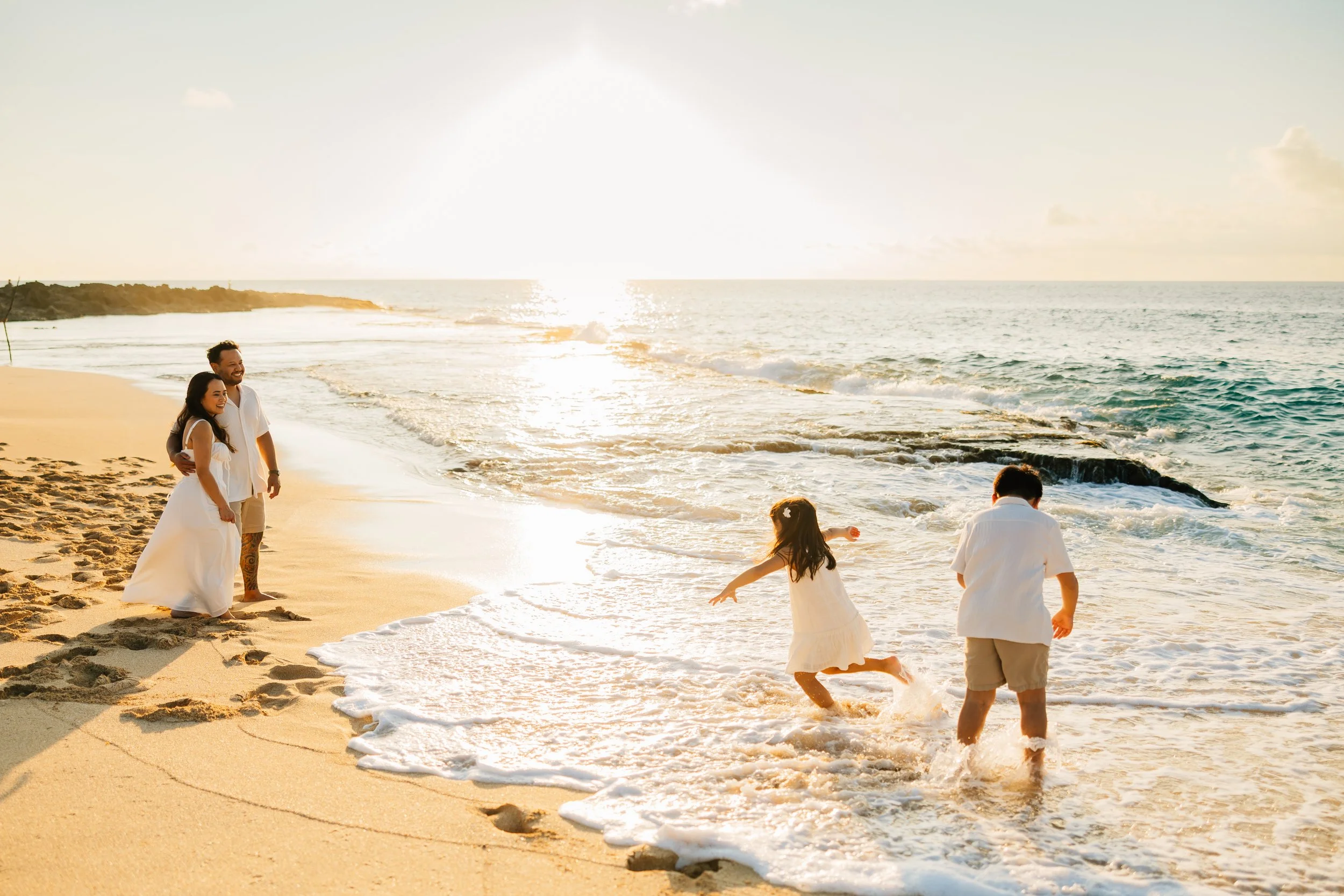 A couple dressed in white, with two children in white dresses, enjoying their time by the ocean at sunset. The woman and man are standing close, smiling, while the children play in the shallow water near the shore.