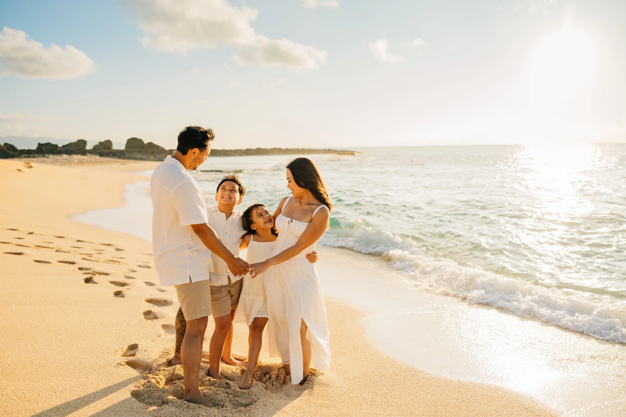 A family of four holding hands on a sandy beach, with the ocean and a blue sky with clouds in the background.