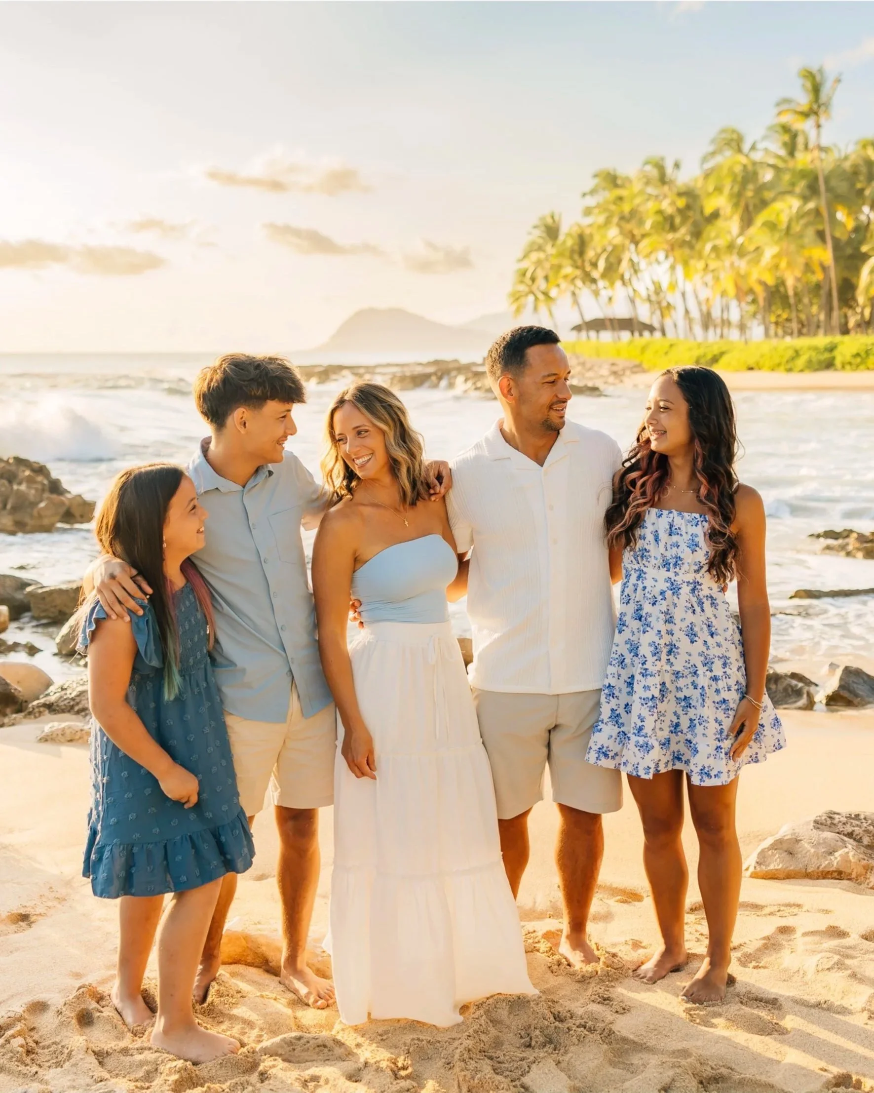 A group of six people, five women and one man, standing arm-in-arm on a sandy beach during sunset, with palm trees and islands in the background, smiling and enjoying each other's company.