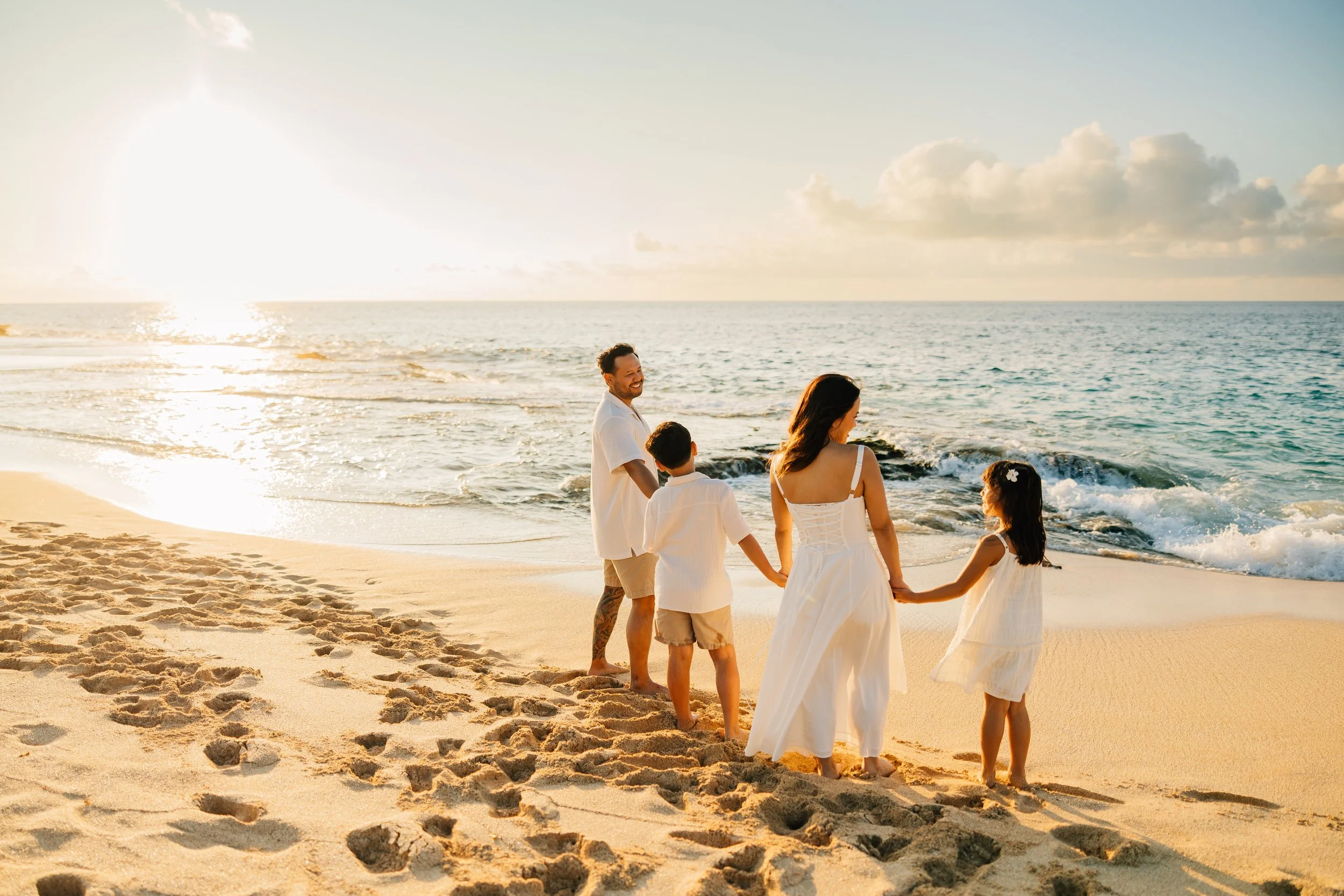 A family of five holding hands and walking on a sandy beach during sunset, with the ocean and sky in the background.