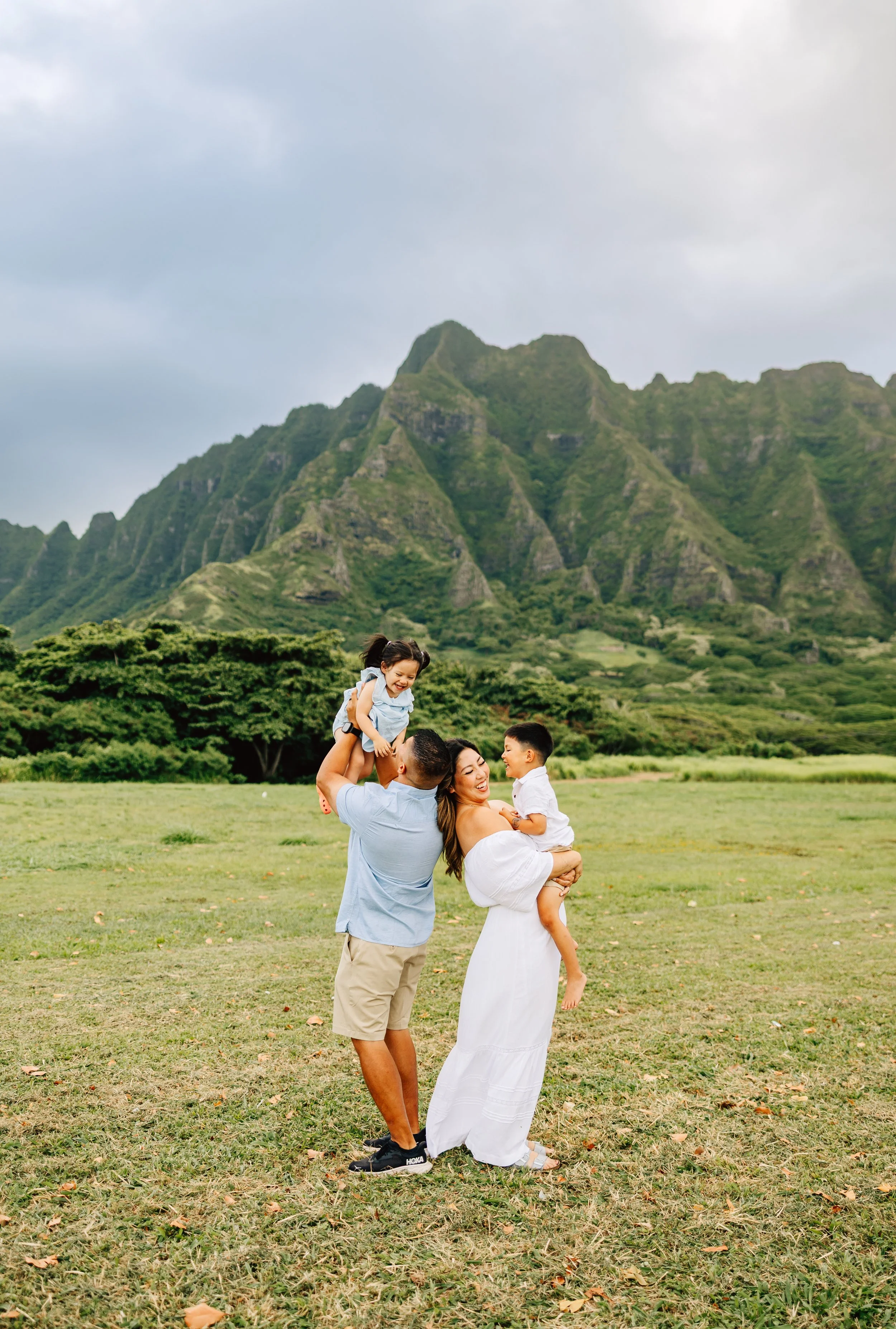 A joyful family of four playing outdoors on a grassy field with green mountain terrain in the background. The father lifts a young girl on his shoulders. The mother, in a white dress, holds a boy in her arms.