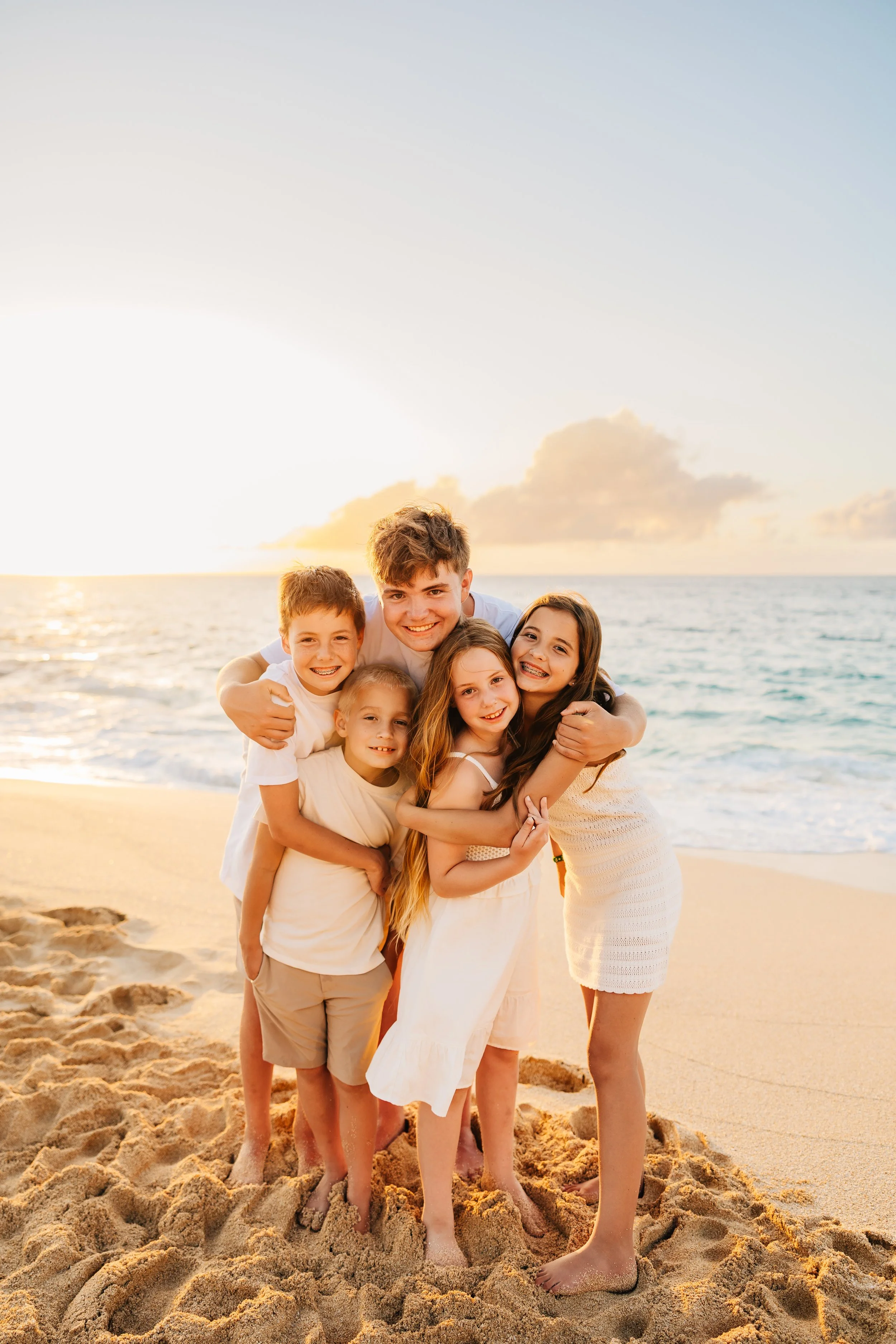 A group of seven children hugging on a sandy beach at sunset, with ocean waves and a partly cloudy sky in the background.