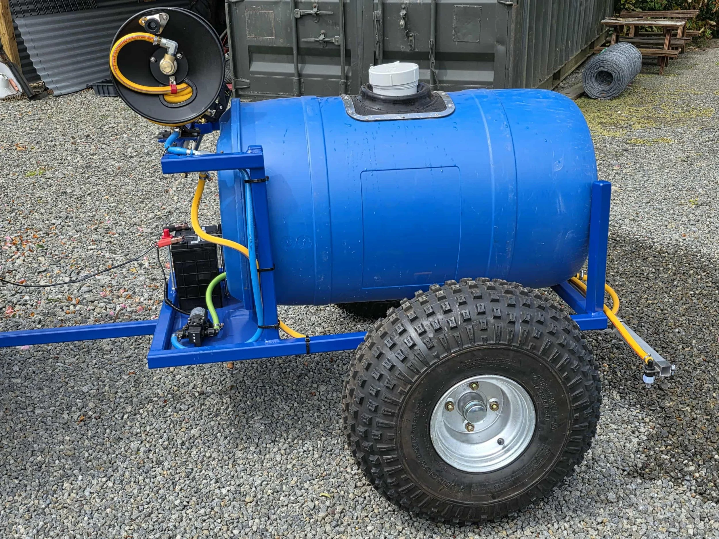 A portable pressure washer with a blue cylindrical tank mounted on a red metal frame, large rugged tires, and various hoses and connections, set outdoors on gravel.