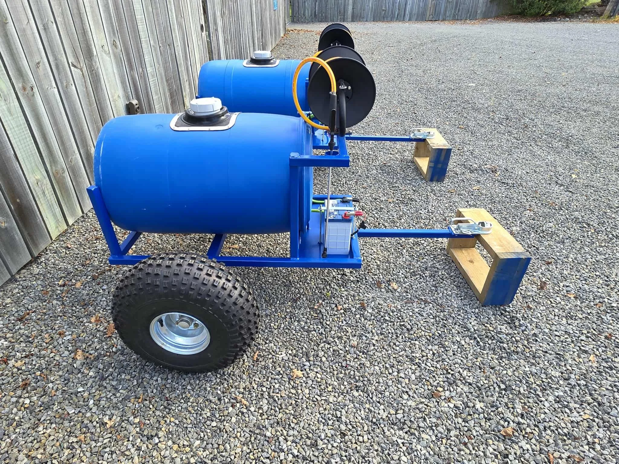 A mobile water tank trailer with two large blue tanks, black hoses, wooden blocks for stabilizing, and large off-road tires, stationed on gravel next to a wooden fence.