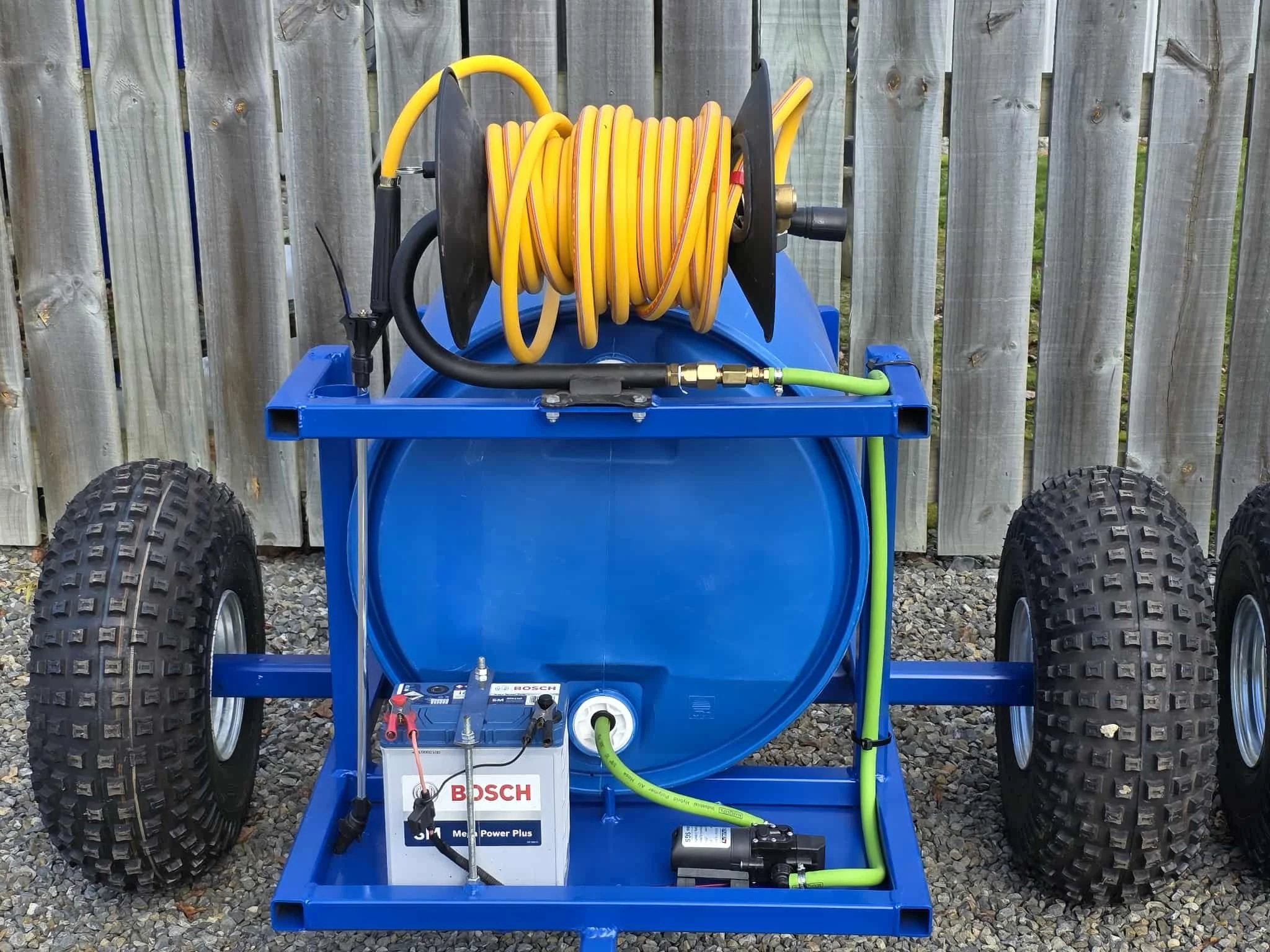 A mobile pressure washer with a blue tank, large tires, a coiled yellow hose on a reel, and a 12v pump, set against a wooden fence and gravel ground.