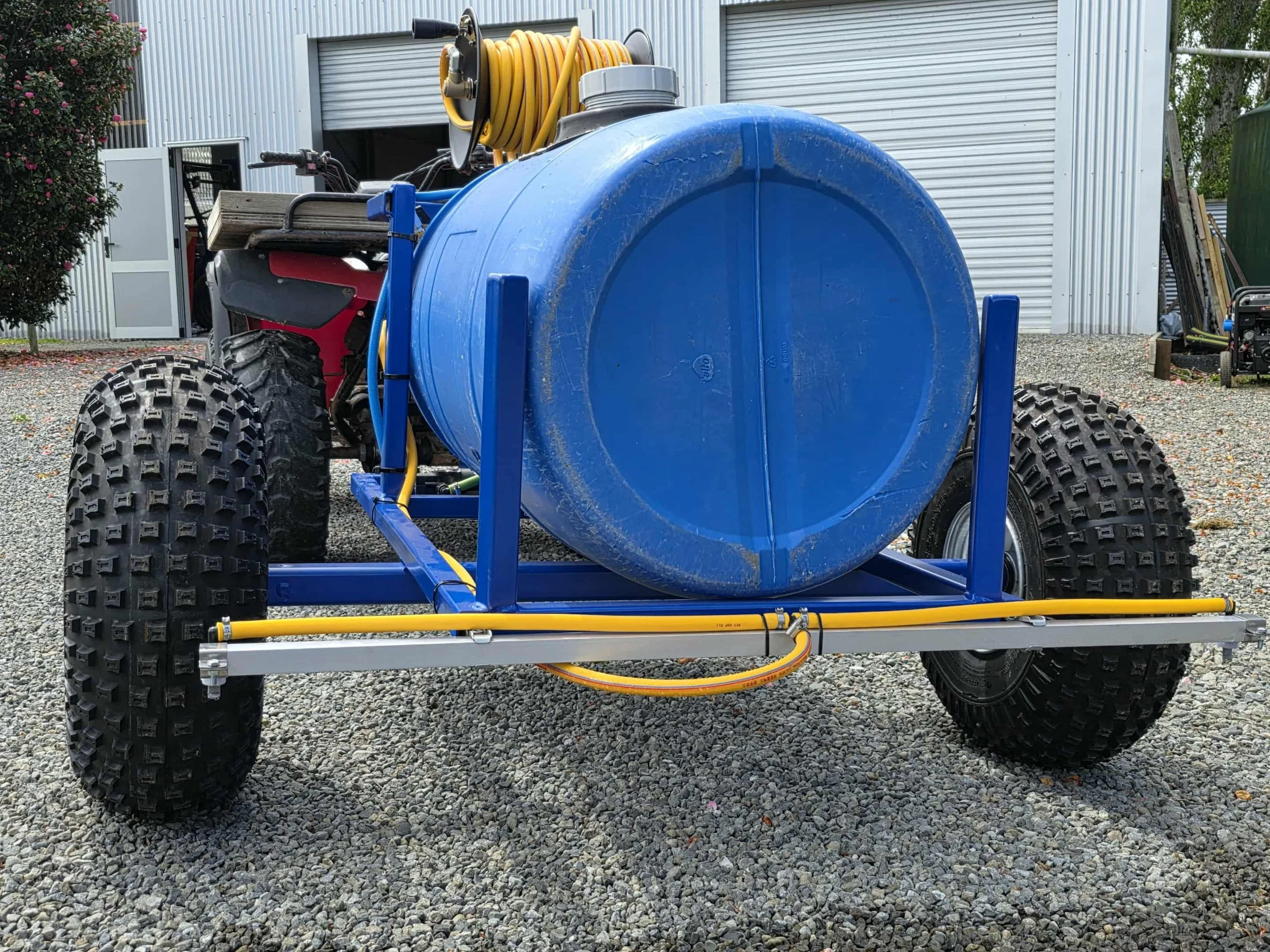 A large blue tank mounted on a blue metal frame with large off-road tires on gravel surface, and a yellow coiled hose on top, in front of a metal building.