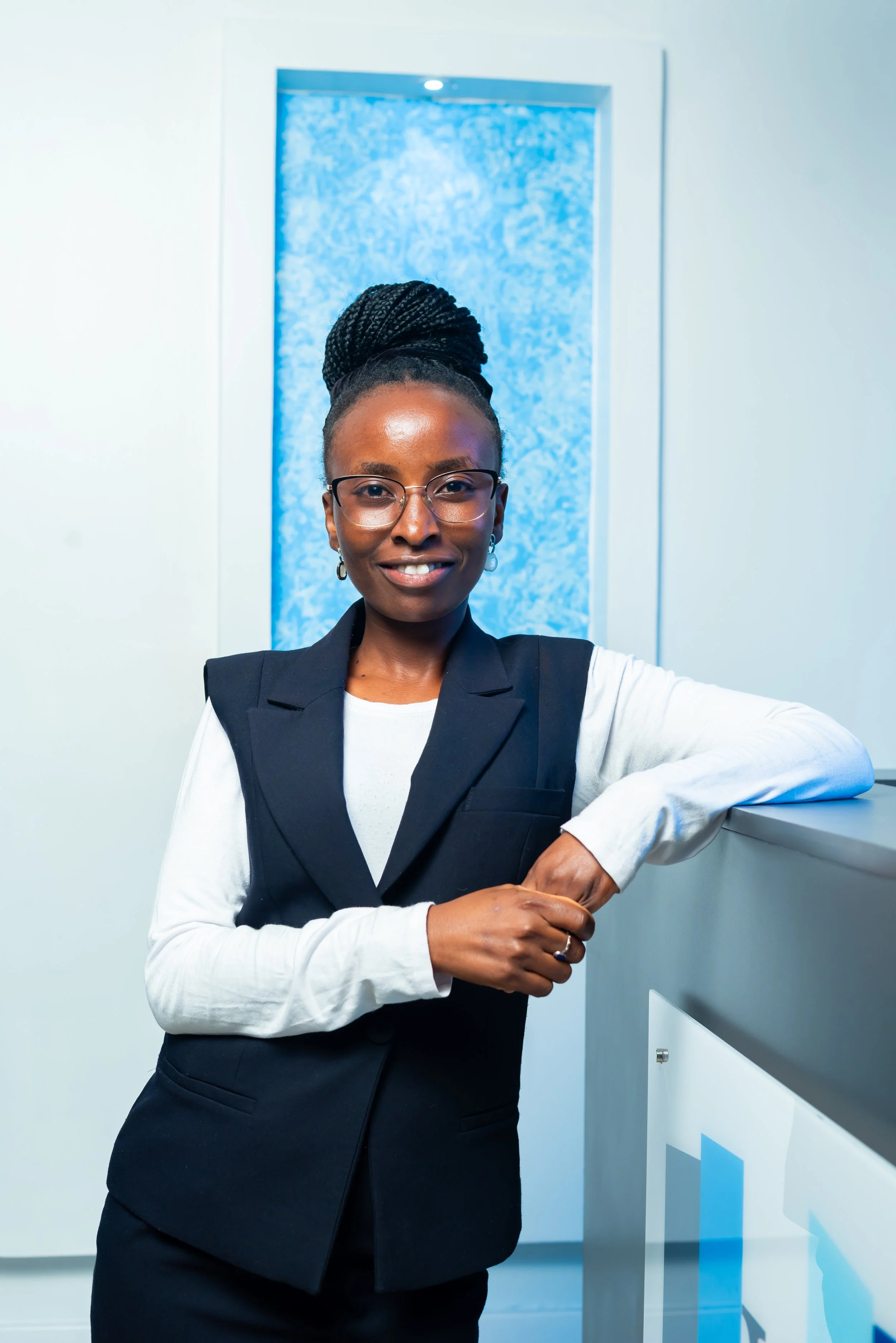 A smiling woman with glasses and braided hair in an updo, wearing a black vest over a white shirt, standing indoors against a blue-lit textured wall and leaning on a counter.