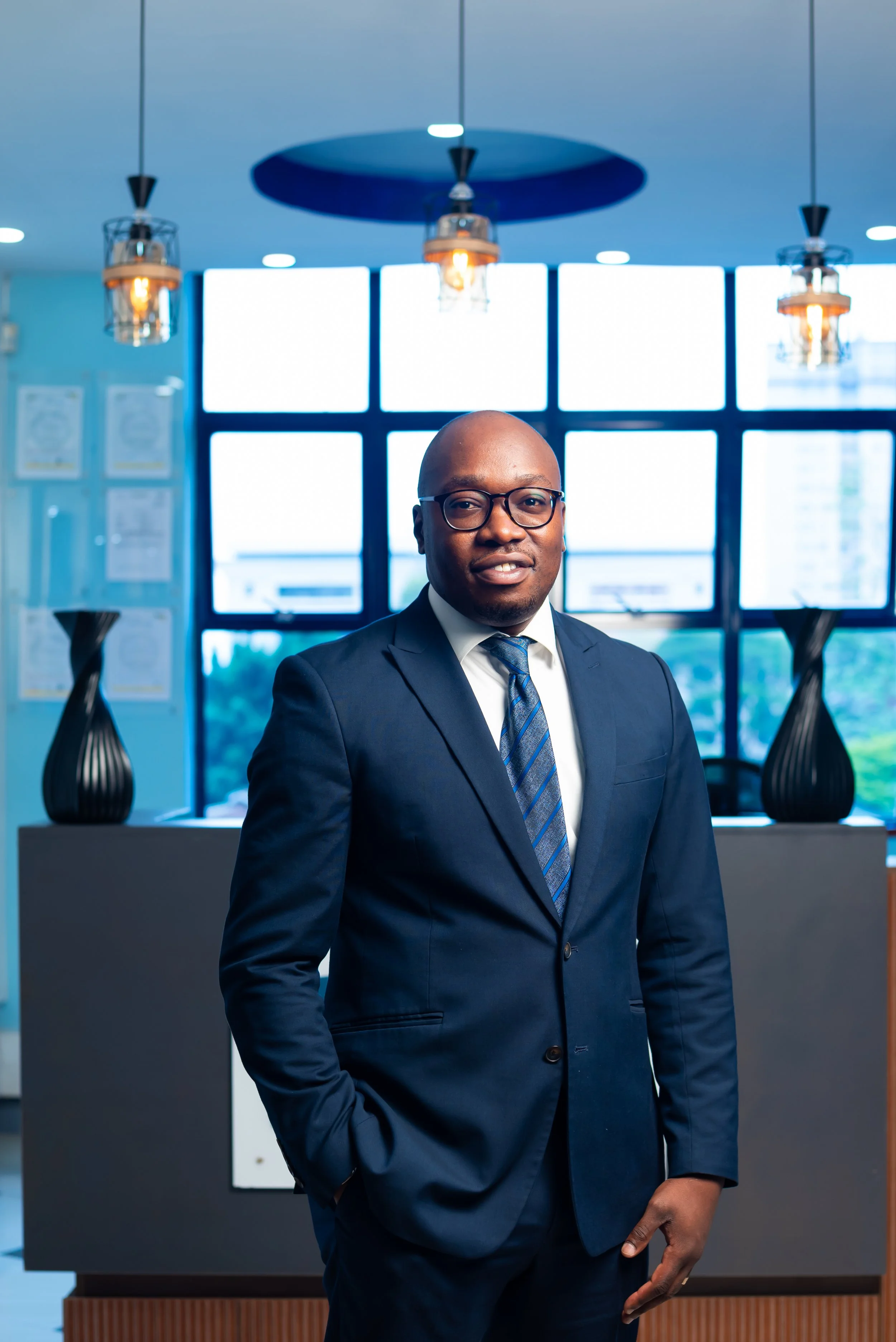 A man in a navy blue suit and glasses standing in an office or lobby area with modern decor and blue accents, including framed artwork and hanging light fixtures.