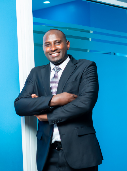 A smiling man in a business suit standing with arms crossed in an office setting with blue walls and glass panels.