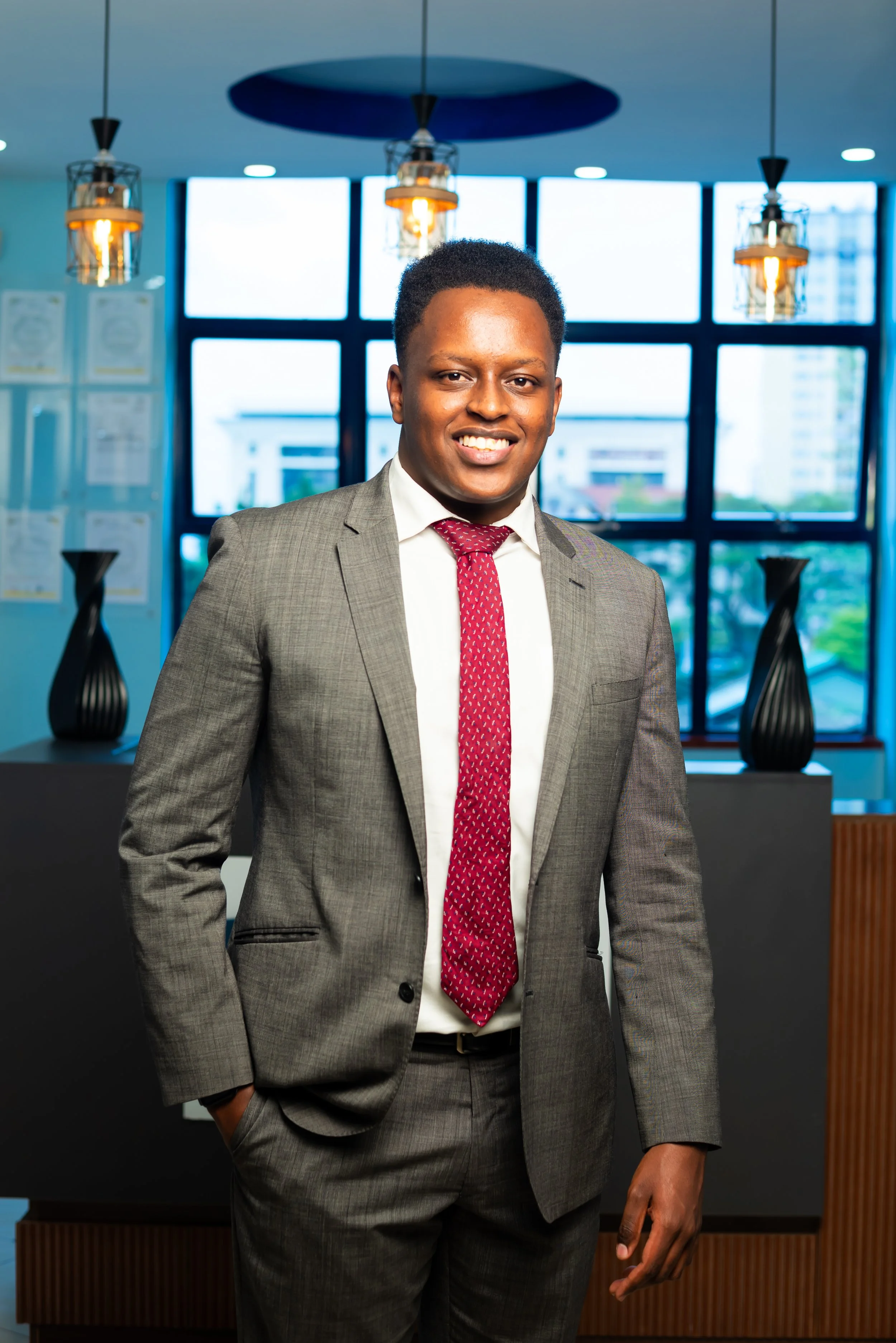 A smiling man in a gray suit, white shirt, and red tie standing in a modern office with large windows and decorative lamps.