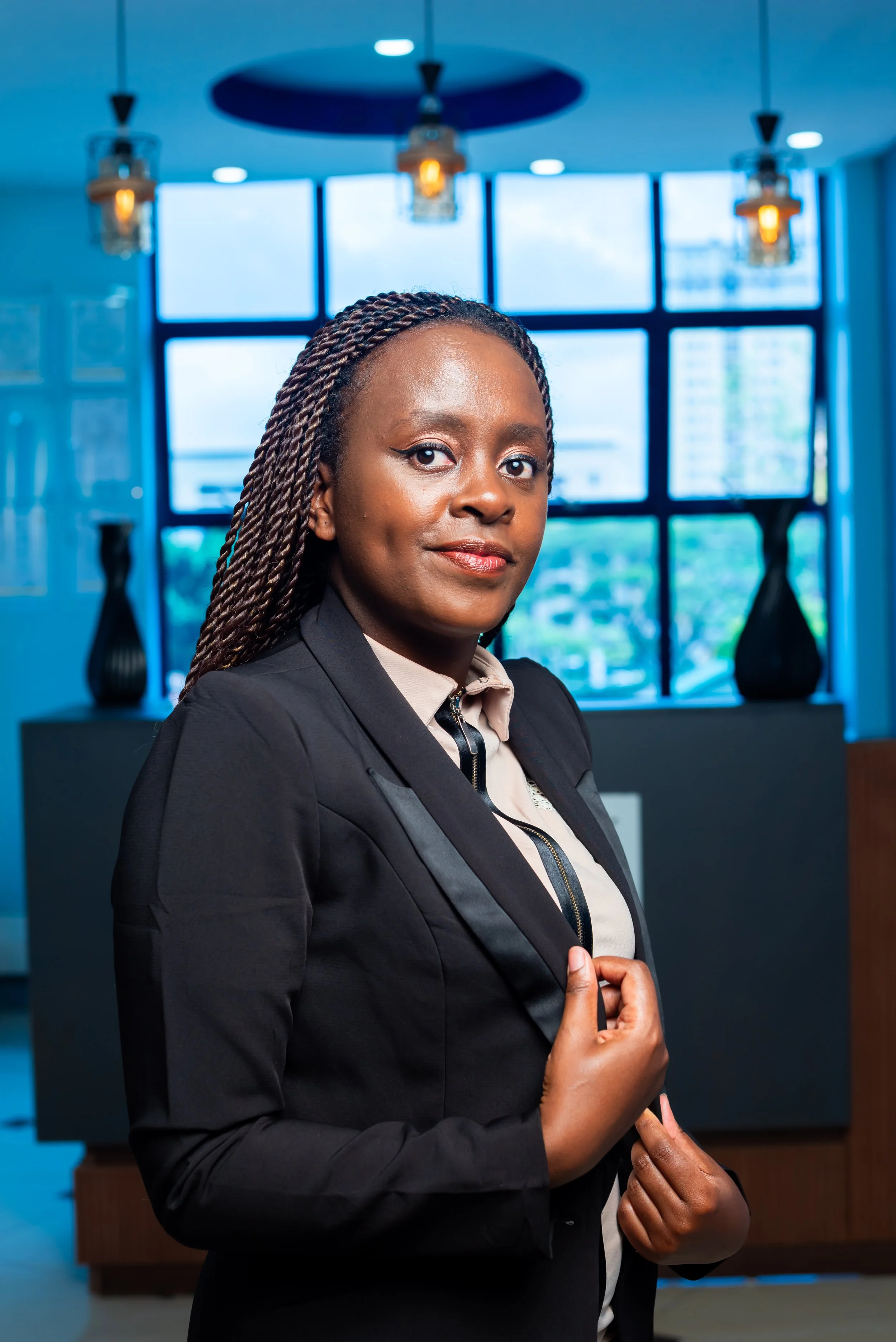A confident woman in business attire standing in a modern office with large windows and decorative vases.