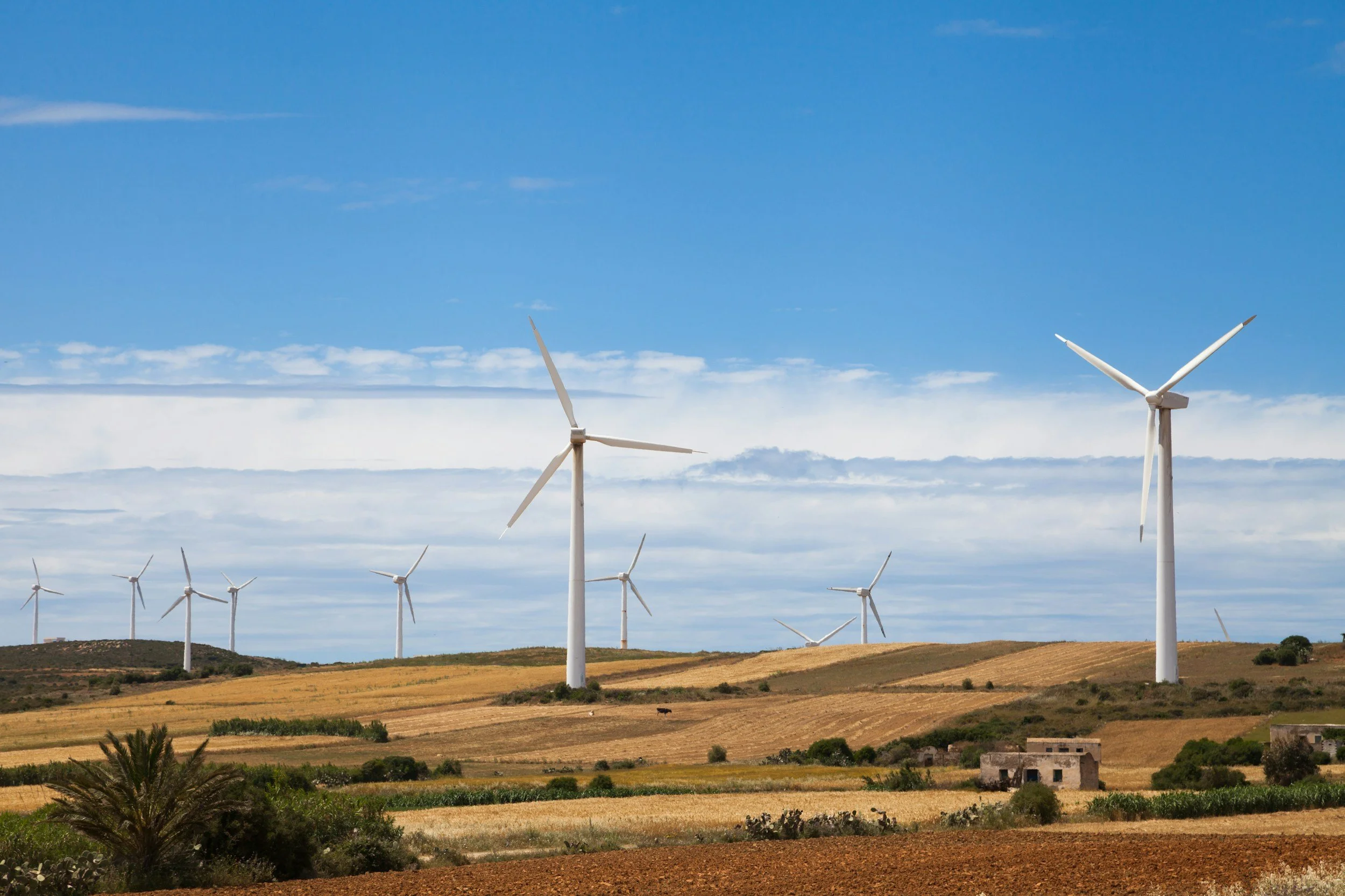 Wind turbines in a rural landscape with fields, small buildings, and a partly cloudy blue sky.
