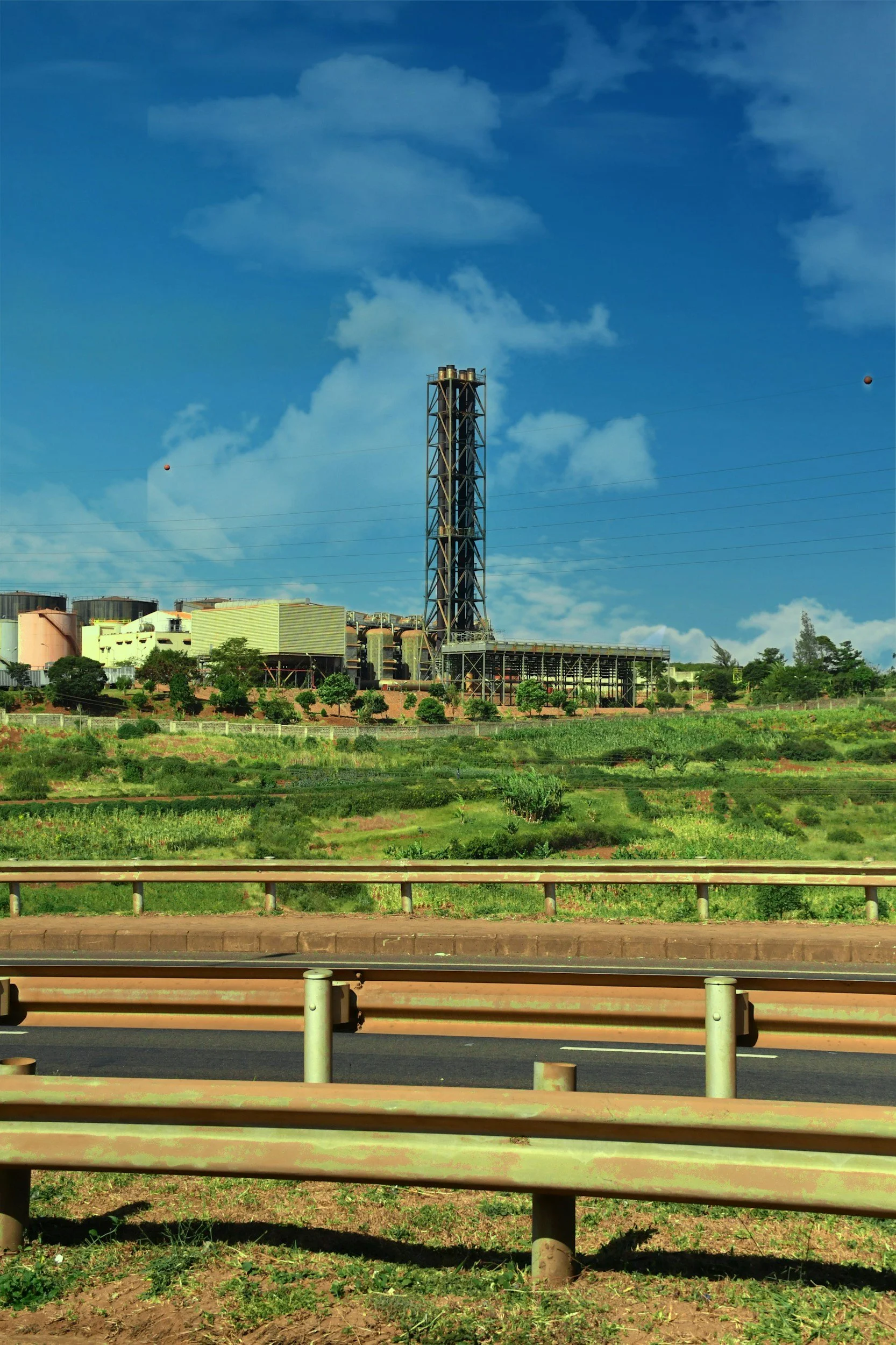 Image of an industrial plant with a tall chimney, situated behind green fields, under a partly cloudy blue sky, viewed from a highway with metal guardrails in the foreground.