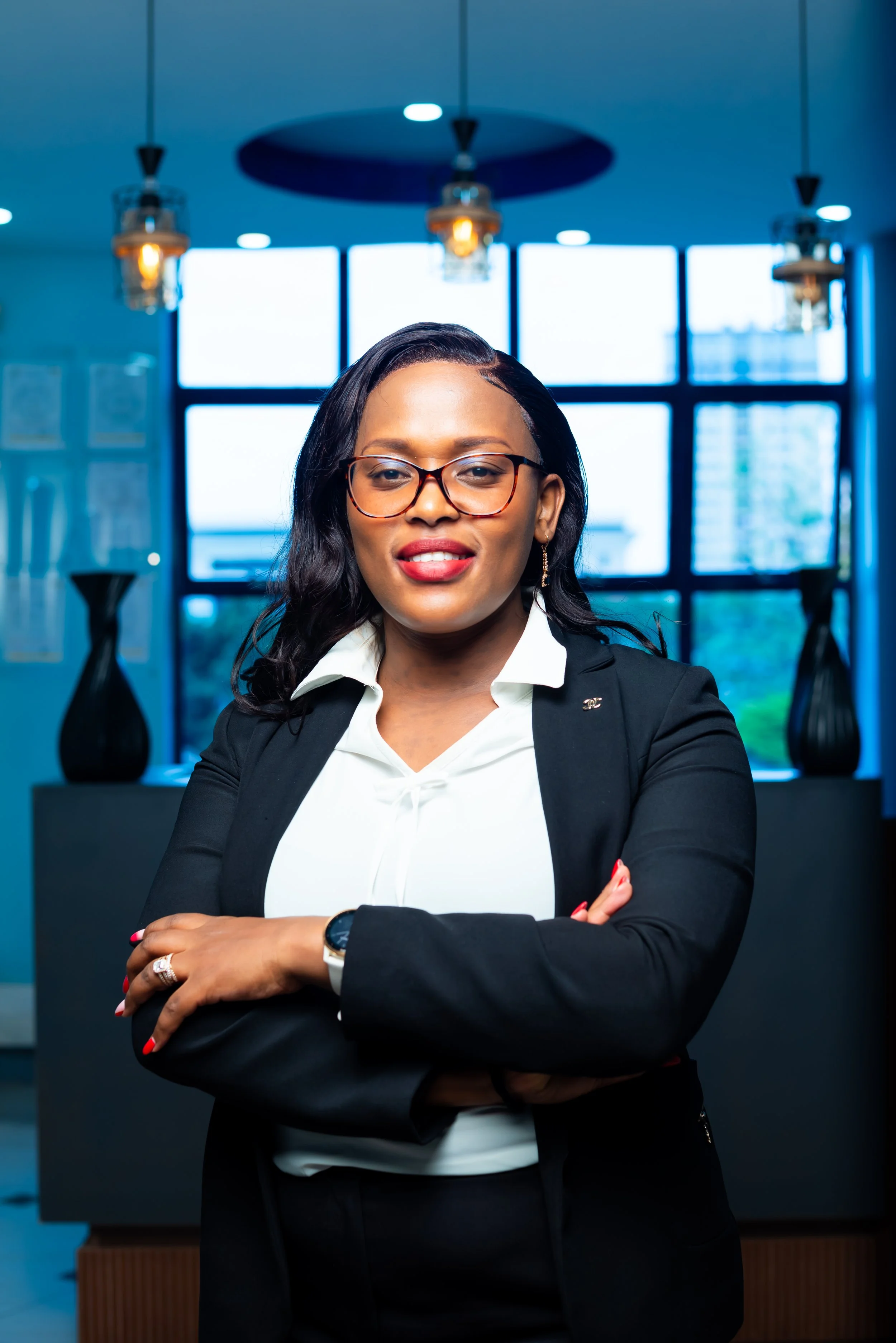 Businesswoman in black blazer and white shirt, wearing glasses, standing in a modern office with large windows, pendant lights, and decorative vases.