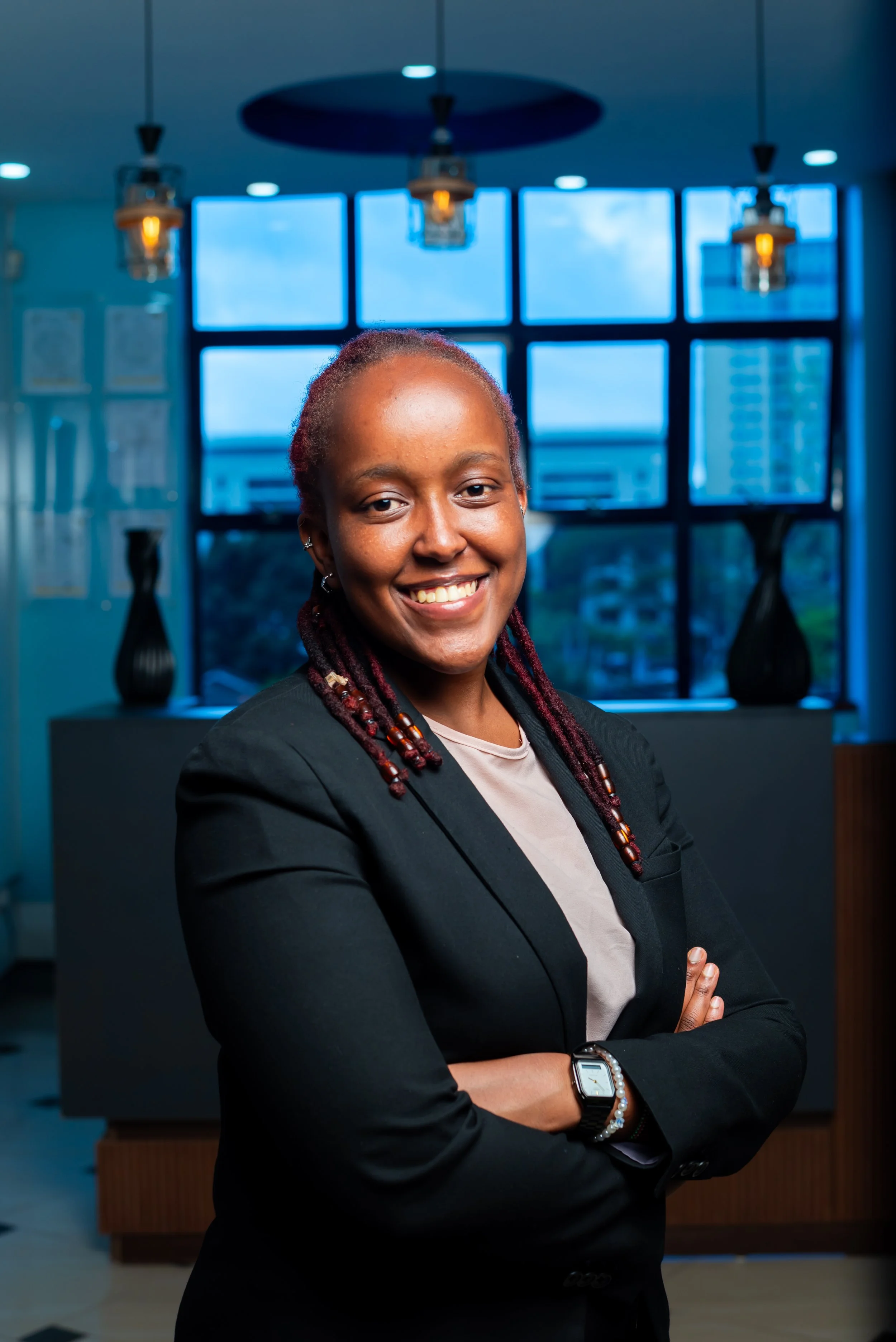 A smiling woman with her arms crossed, wearing a black blazer and a smartwatch, standing in an office with large windows and cityscape views.