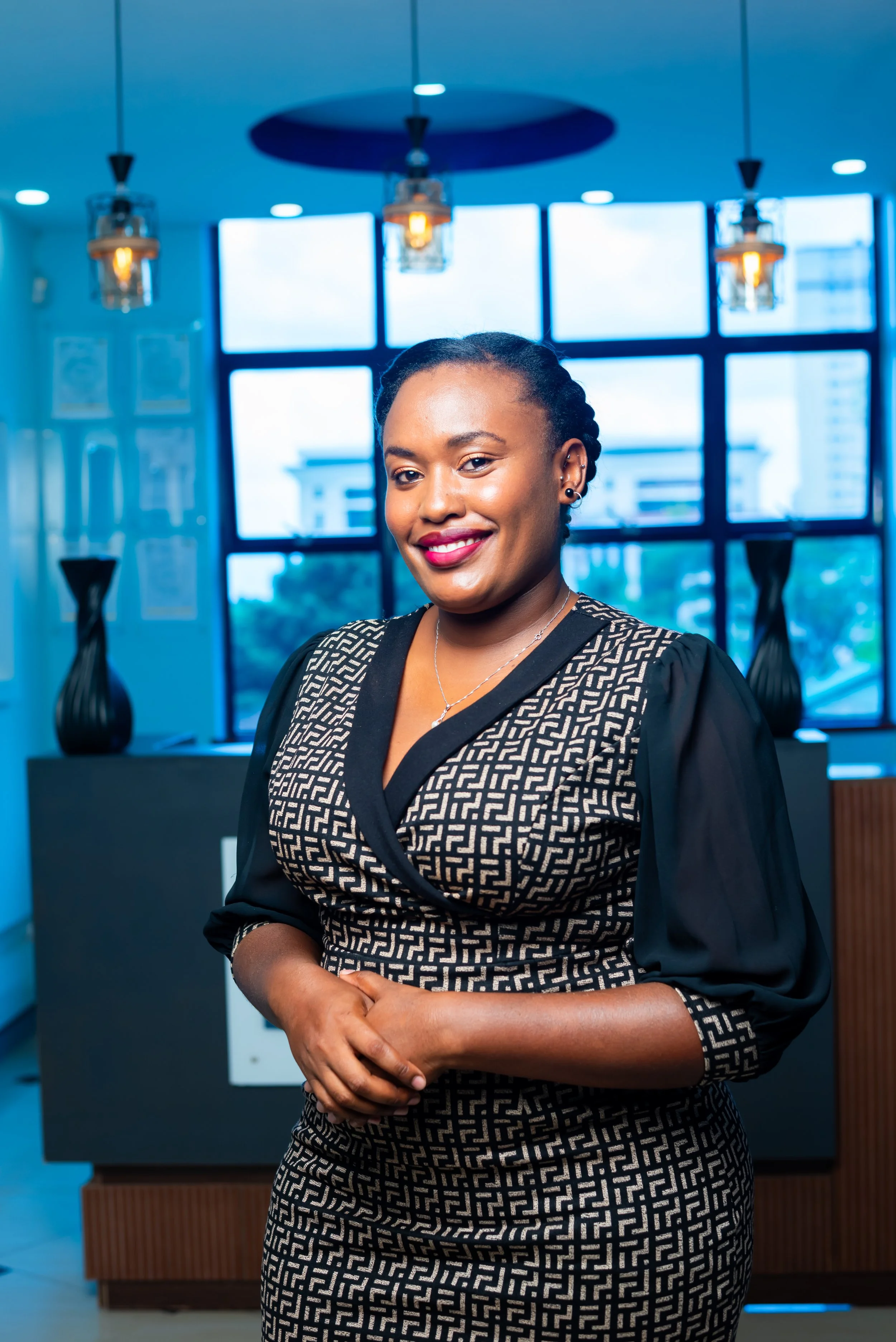 A smiling woman with dark hair pulled back, wearing a black and beige patterned dress, standing in a modern office with large windows and decorative vases.