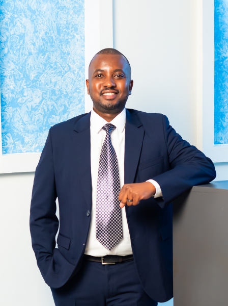 Professional man in navy suit and tie smiling, standing with arm resting on a desk in a bright office environment.