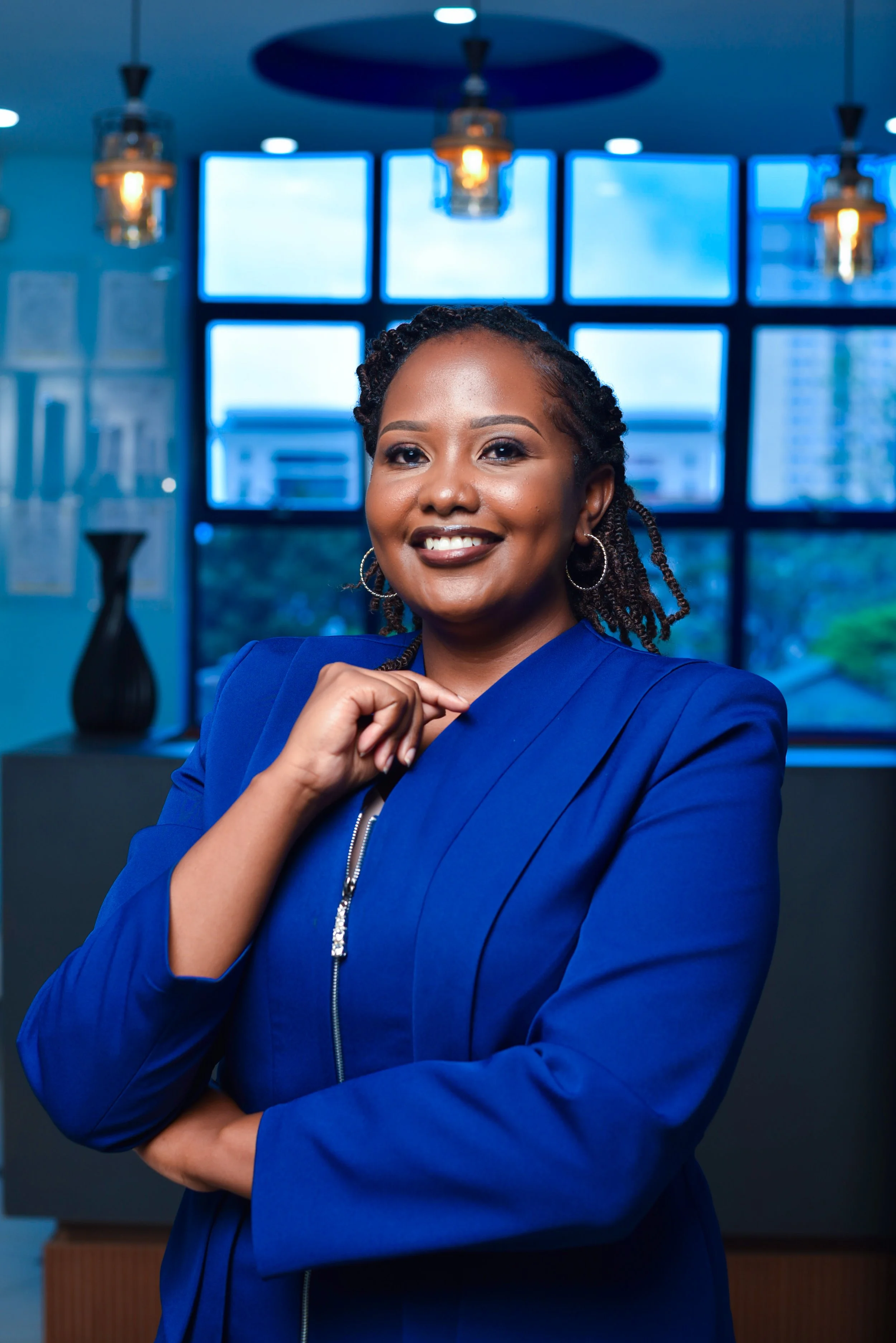 Portrait of a smiling woman with braided hair wearing a blue blazer in an office setting with large windows and decorative lighting.