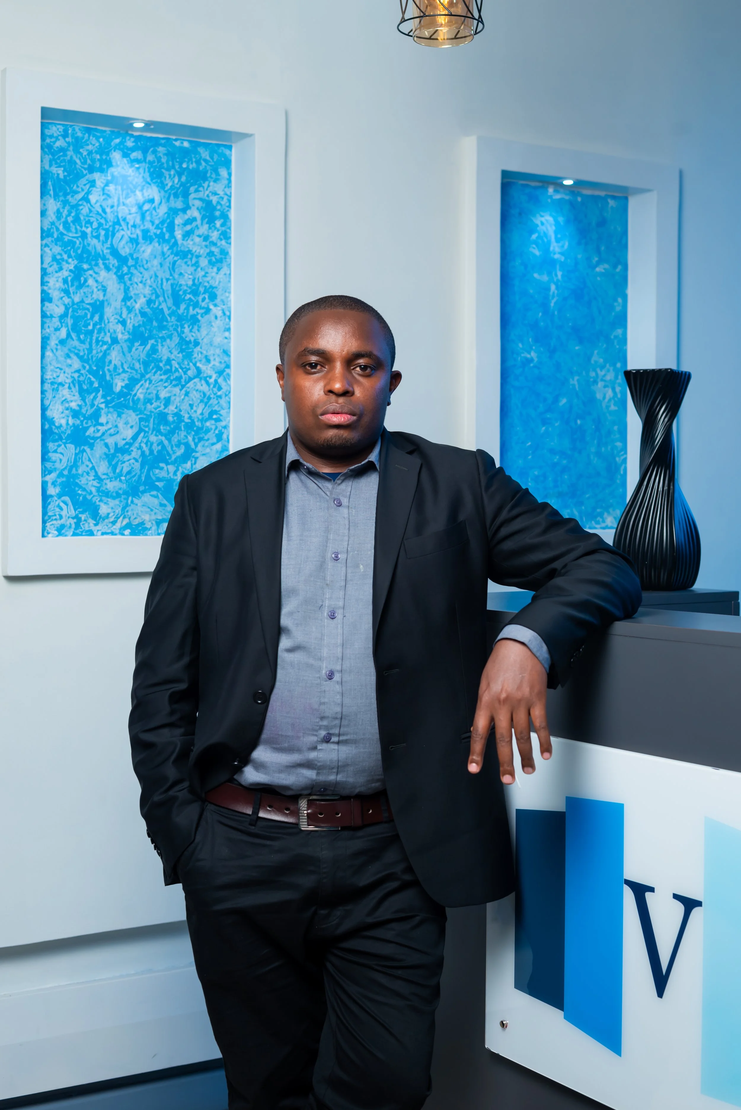 A man in a dark suit and gray shirt standing in an office or lobby area with blue abstract artwork on the wall and a modern black vase on a counter.