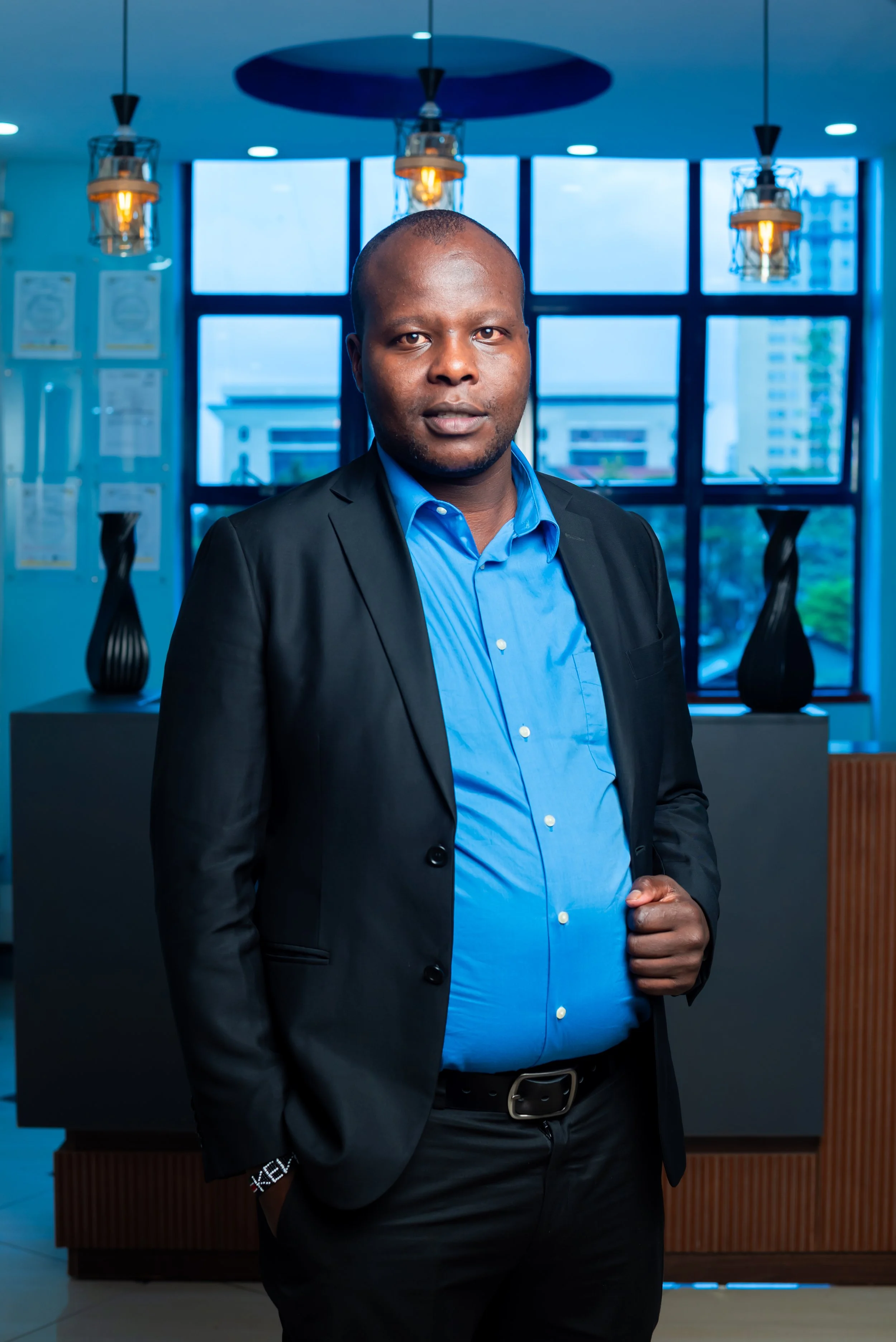 A man in a black suit and blue dress shirt standing in a modern office or hotel lobby with large windows, hanging lights, and decorative vases.