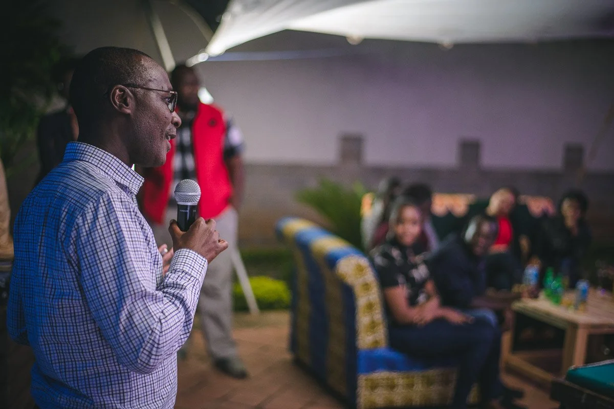 A man with glasses and a checkered shirt speaks into a microphone at an outdoor gathering, with a group of seated people listening in the background.