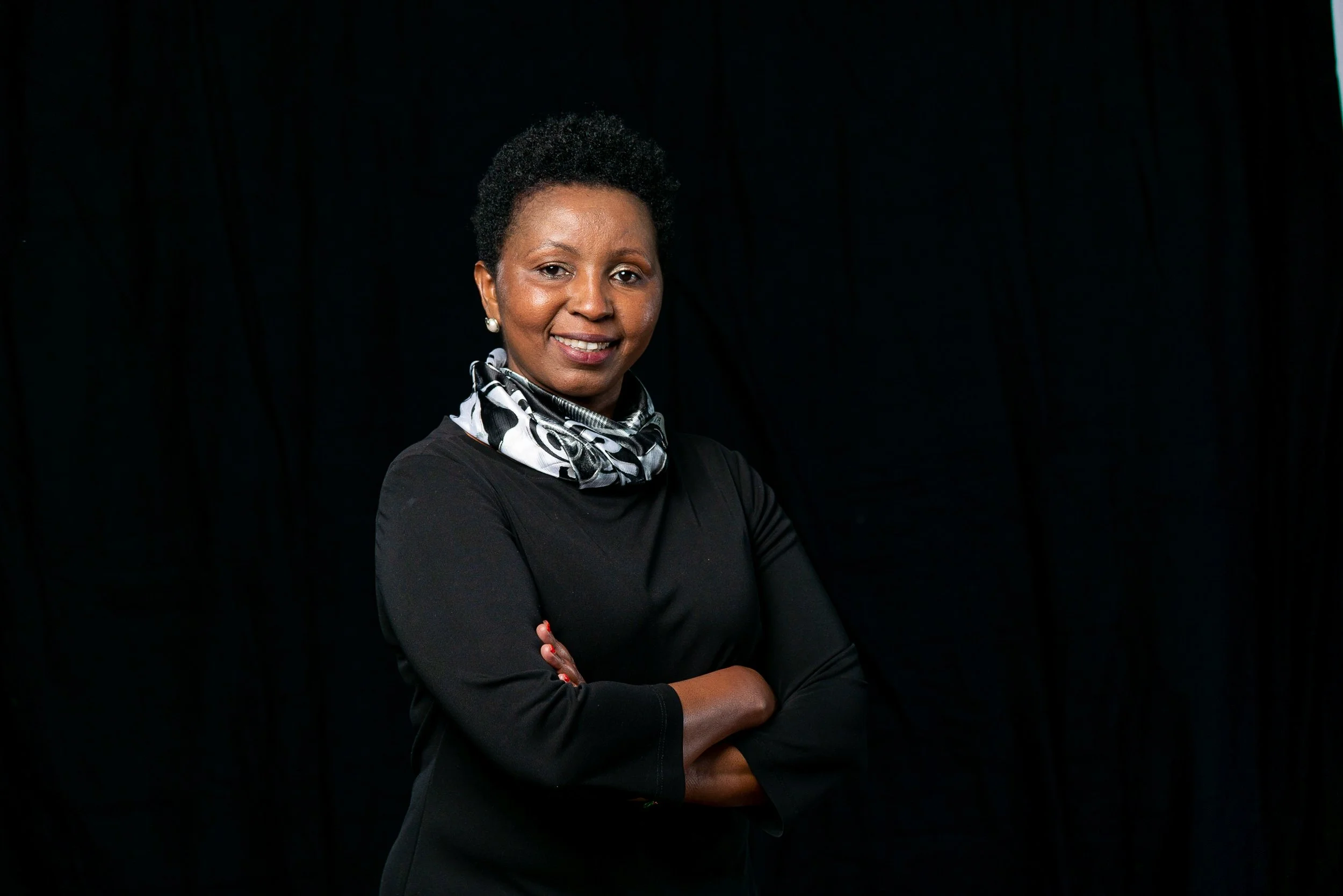 A woman with short curly dark hair and glasses, dressed in a houndstooth patterned jacket and scarf, standing indoors with large windows and modern decor in the background.