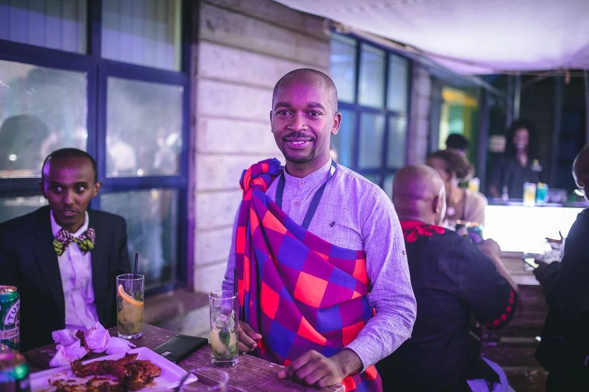 Emmanuel Laalia smiling at a social gathering, wearing a traditional patterned cloth over a gray shirt, seated at a table with drinks and food, with other people in the background.