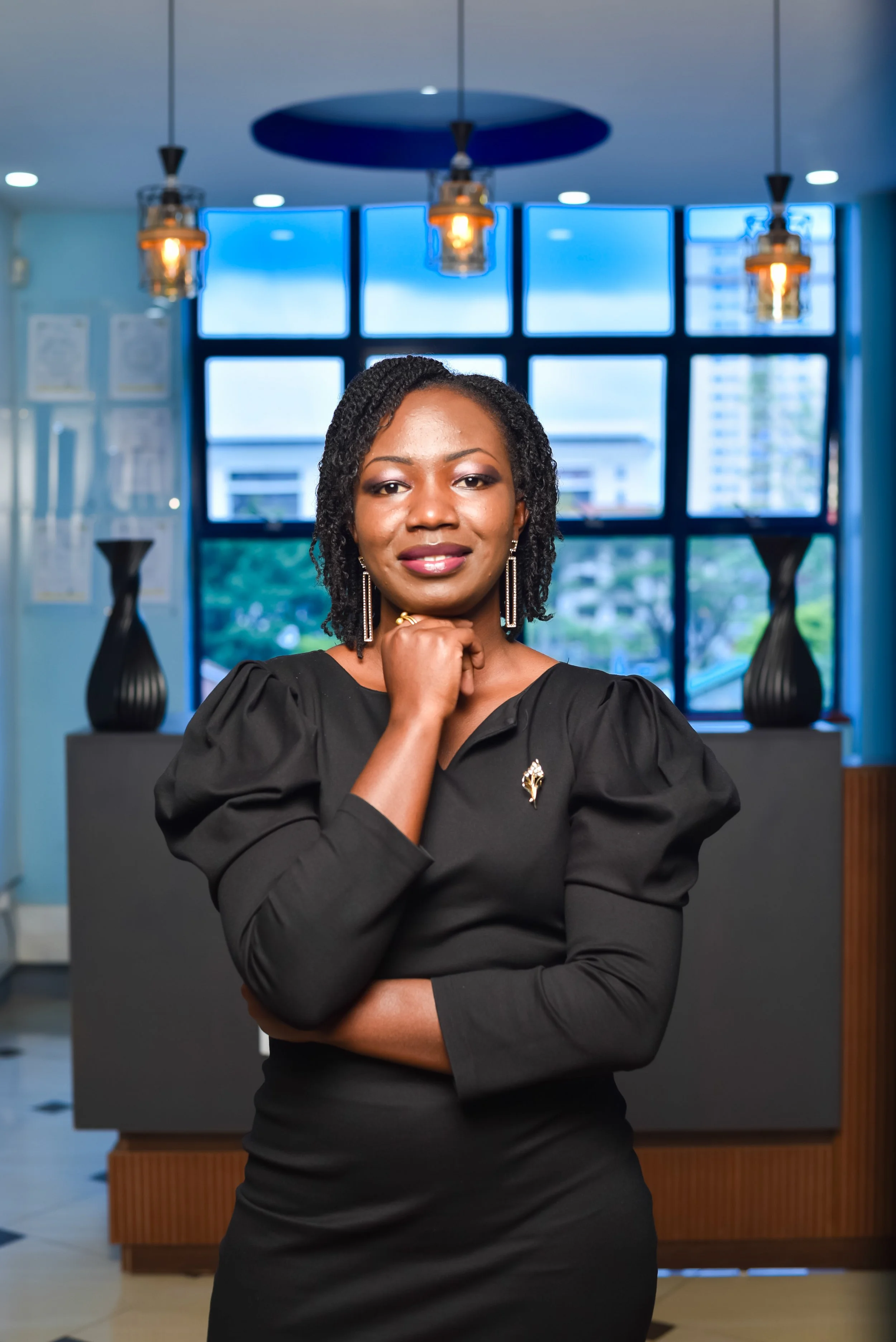 A confident woman in a black dress, standing indoors with a large window behind her, illuminated by hanging pendant lights.