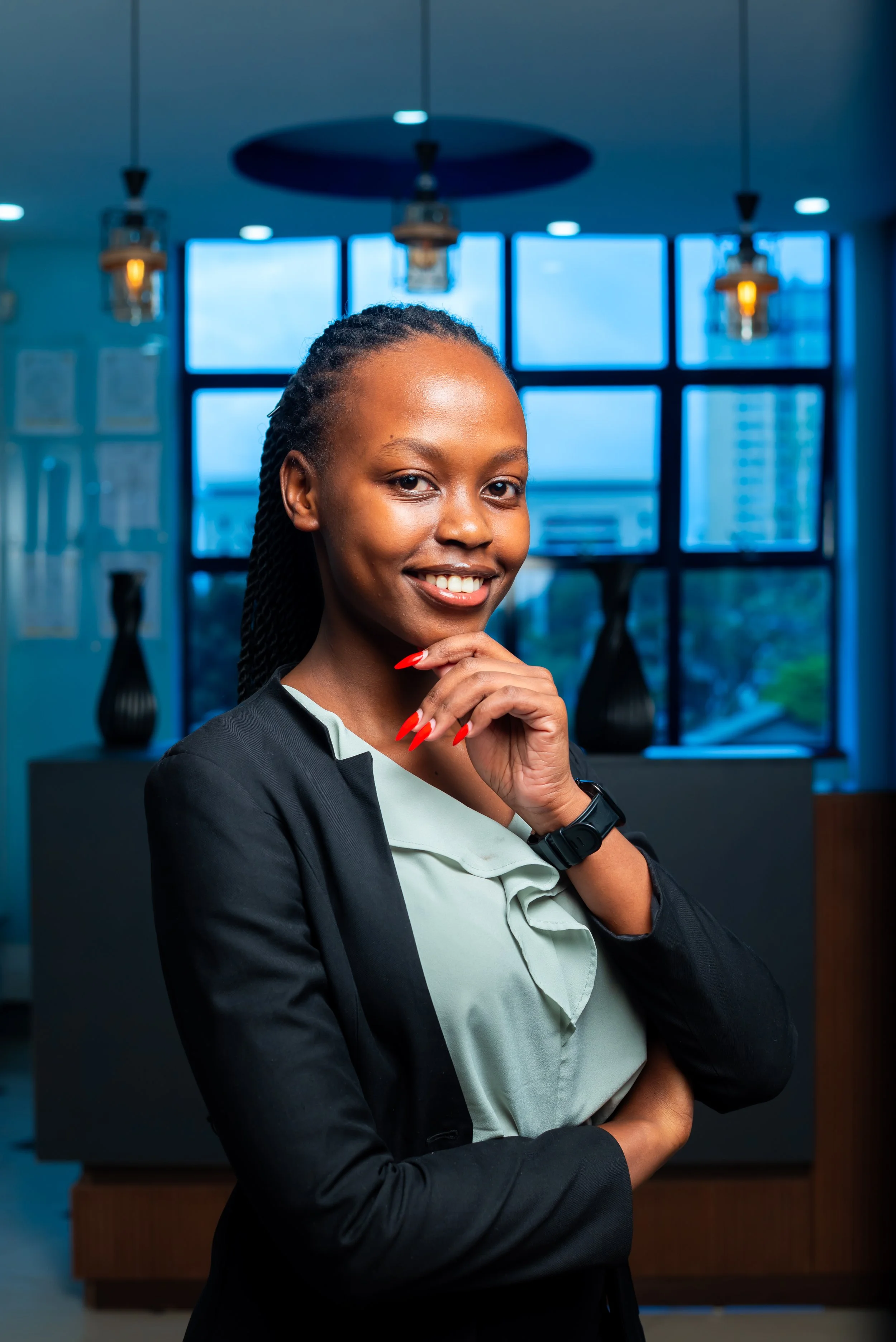 A confident woman in business attire, smiling and posing with hand near chin, in an office with large windows and modern decor.
