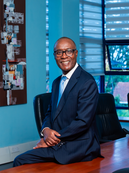 Alex Muriuki, Viva's Senior Partner smiling man in a navy blue suit, glasses, and a blue tie sitting at a wooden desk in an office with large windows and abstract artwork on the wall.
