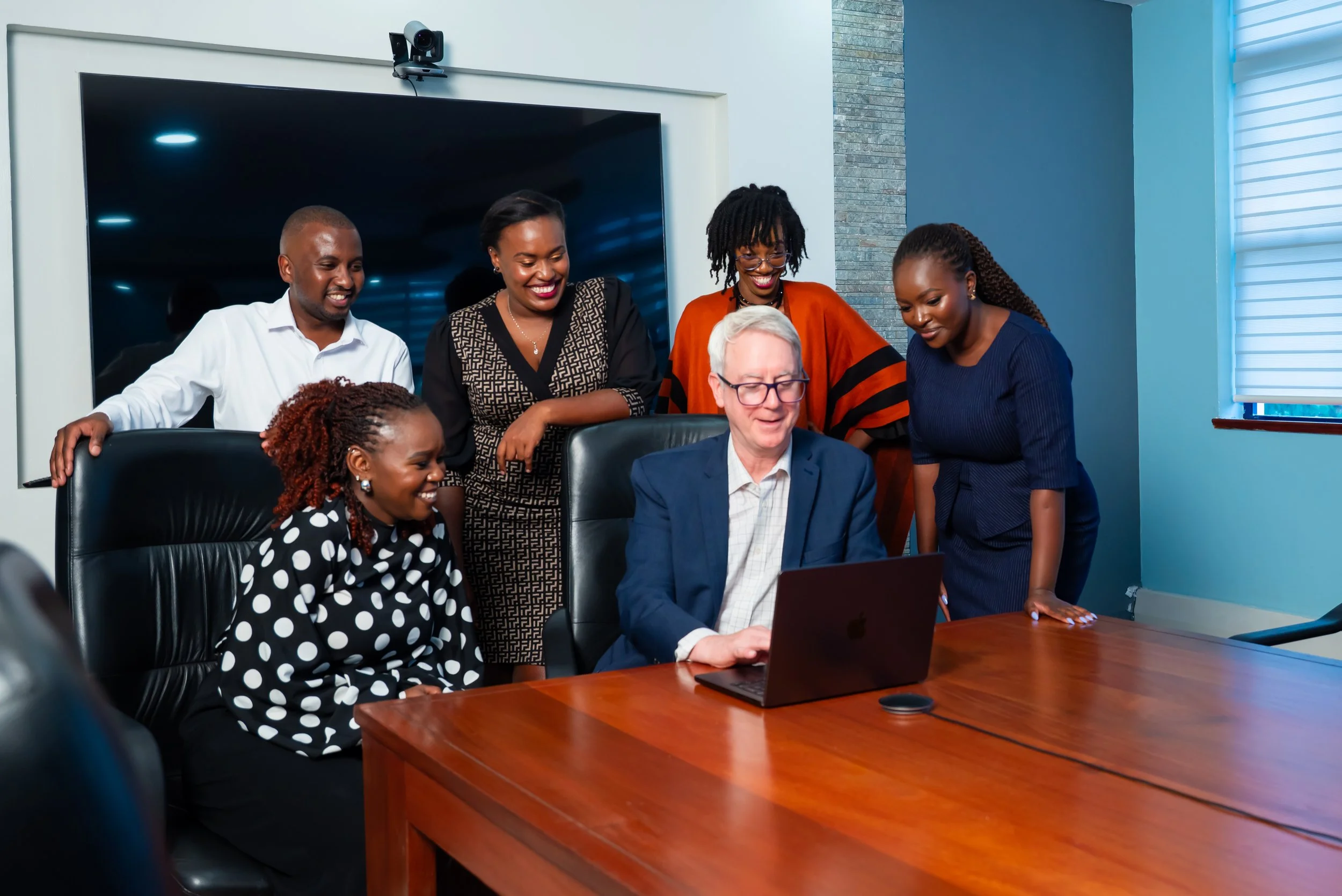 Six Viva team members in a meeting room gathered around a laptop, smiling and looking at the screen. The group includes four women and two men. The room has a large TV or monitor on the wall and a window with blinds.