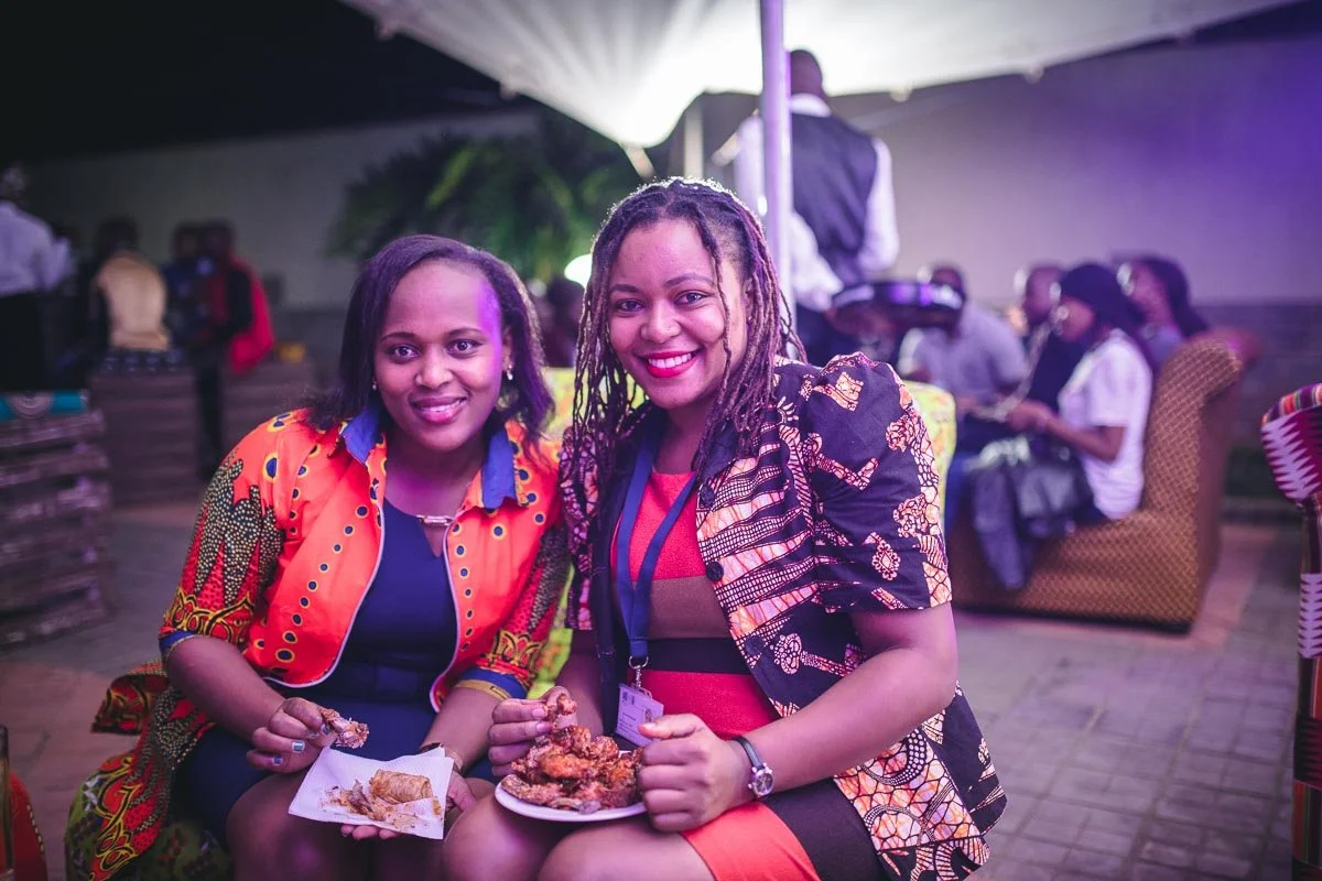 Two women sitting together at an outdoor event, smiling and holding plates of food. There are other people in the background, some sitting and some standing, under a canopy with soft lighting.