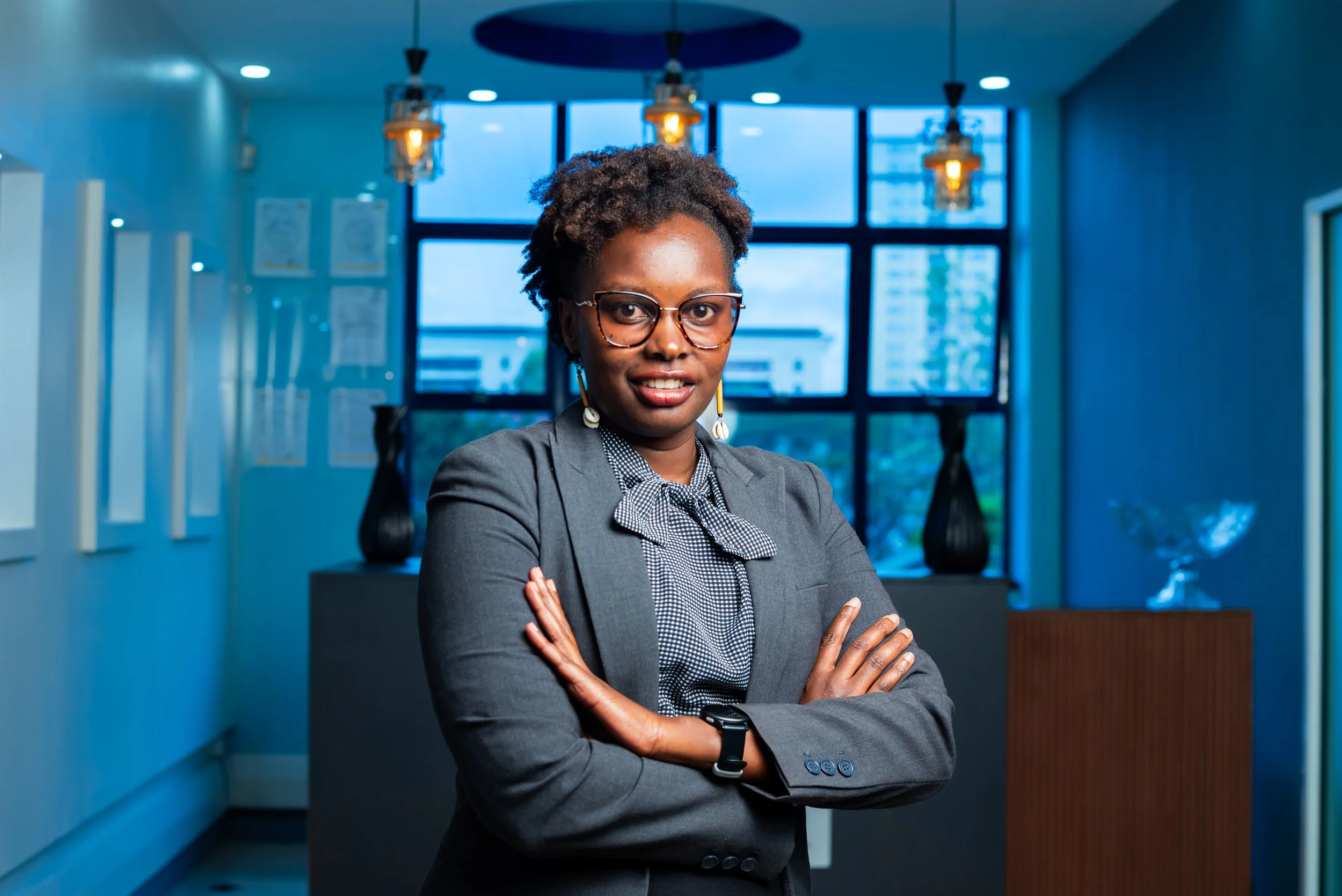 A confident woman with glasses and earrings, wearing a gray blazer and a bow tie blouse, standing with arms crossed in an office with blue lighting and large windows.