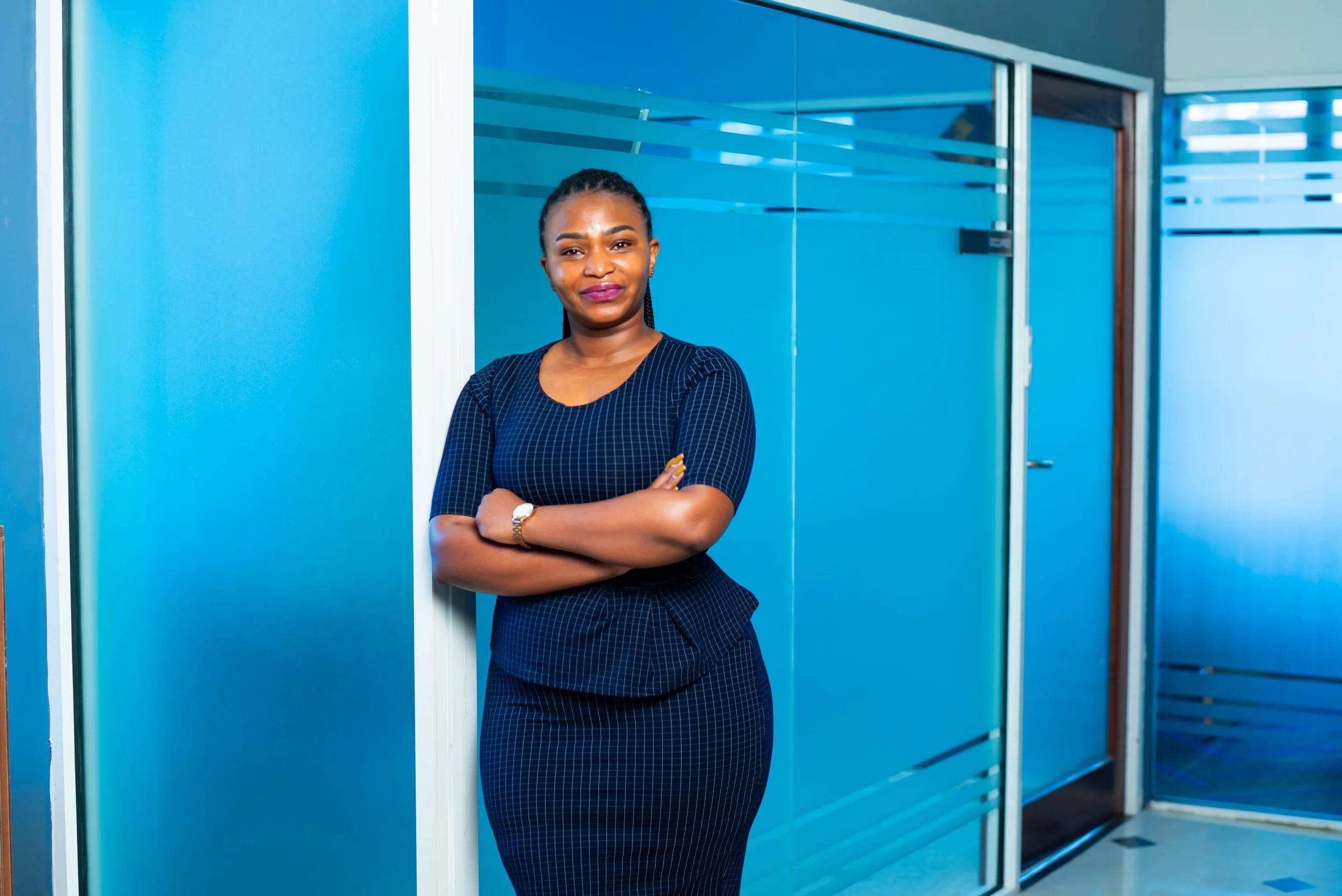 A professional woman in a navy blue checkered dress standing with arms crossed in an office corridor with blue walls and glass doors.