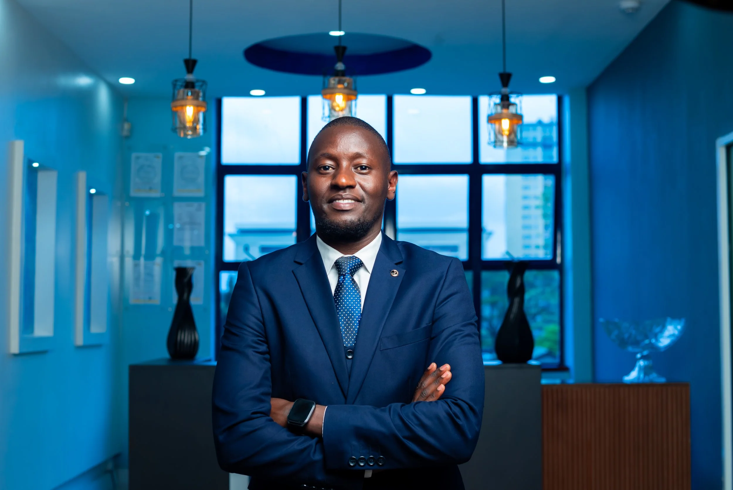 A man in a blue suit standing with arms crossed in an office or modern building lobby with large windows, decorative lighting, and artwork.