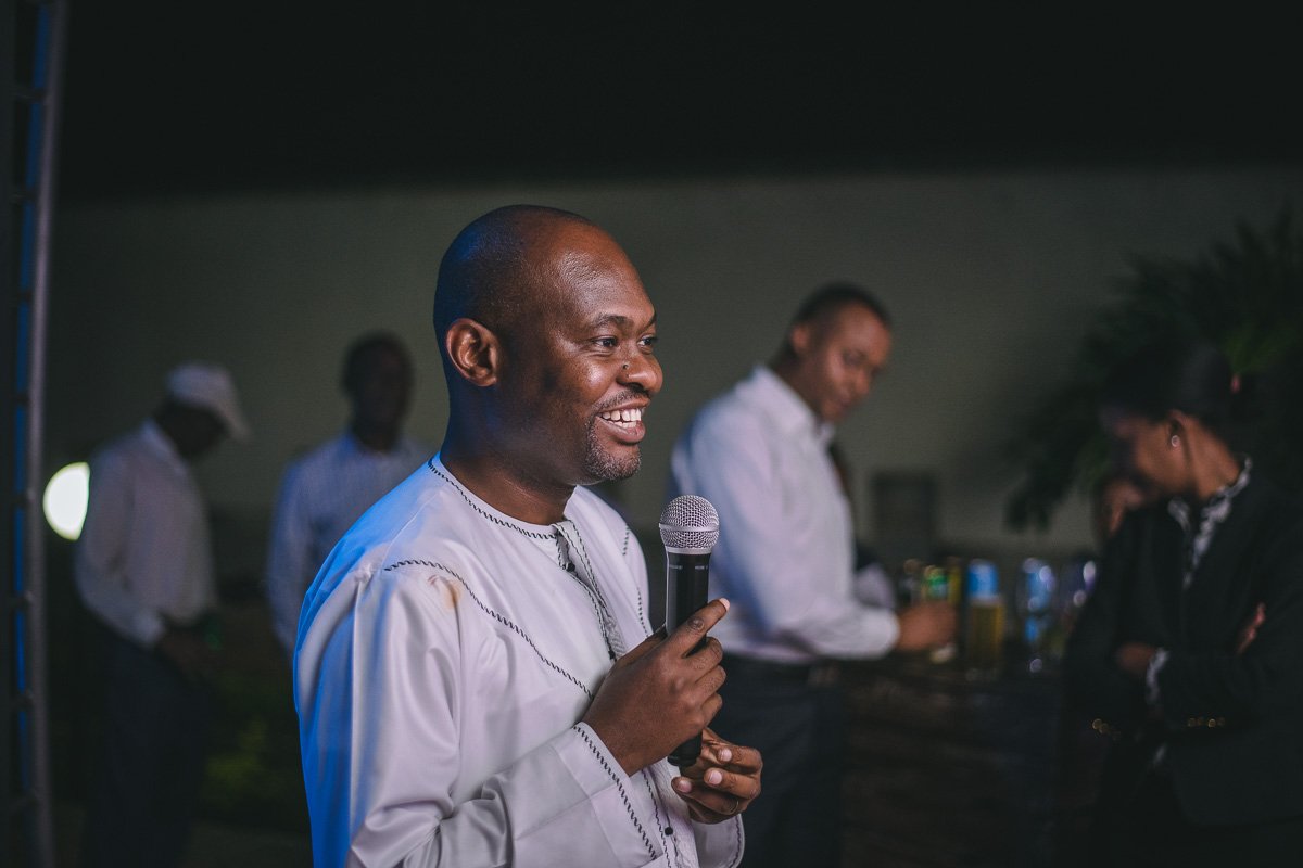  Edward Mwachinga smiling while holding a microphone at an indoor event. Other people are in the background, some looking down or away, with drinks on a table.