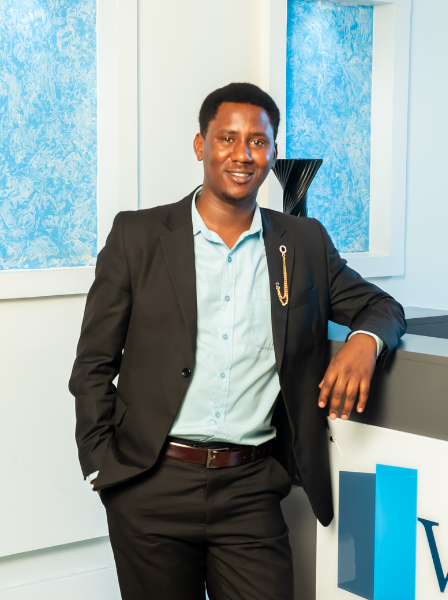 Man in a black suit with a light blue shirt, leaning on a white counter with a blue and white background.