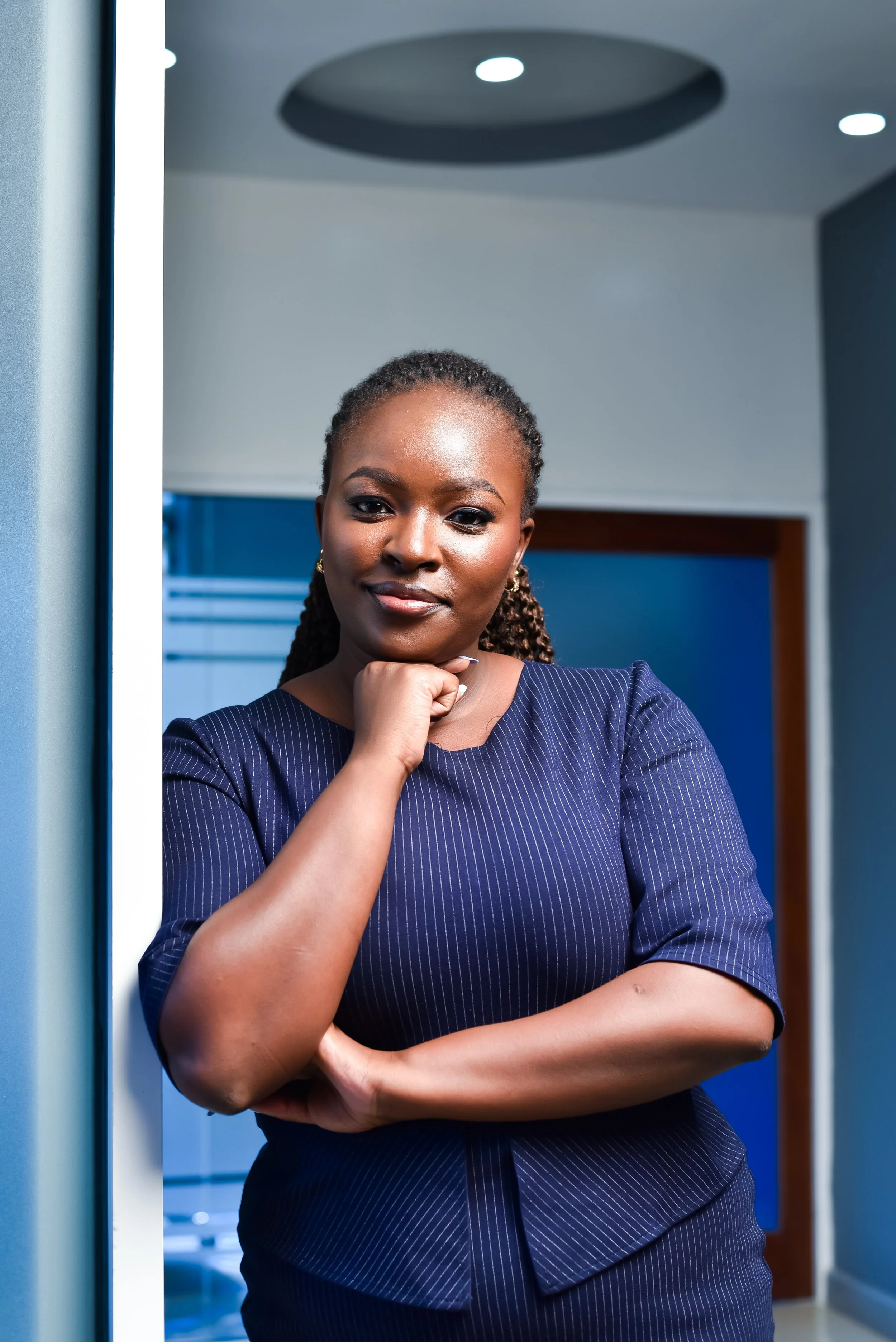 A confident woman with braided hair, wearing a dark pinstriped dress, standing with her chin resting on her hand in a modern office setting.