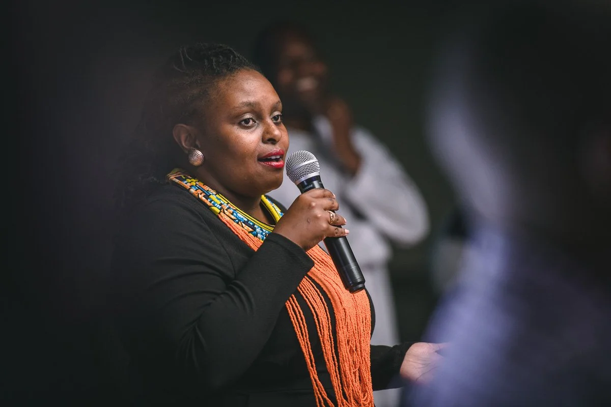 Anne Mubia-Murungi, wearing colorful bead necklaces and earrings, holding a microphone and speaking at an event. She is dressed in a black outfit, and there are women smiling in the background.