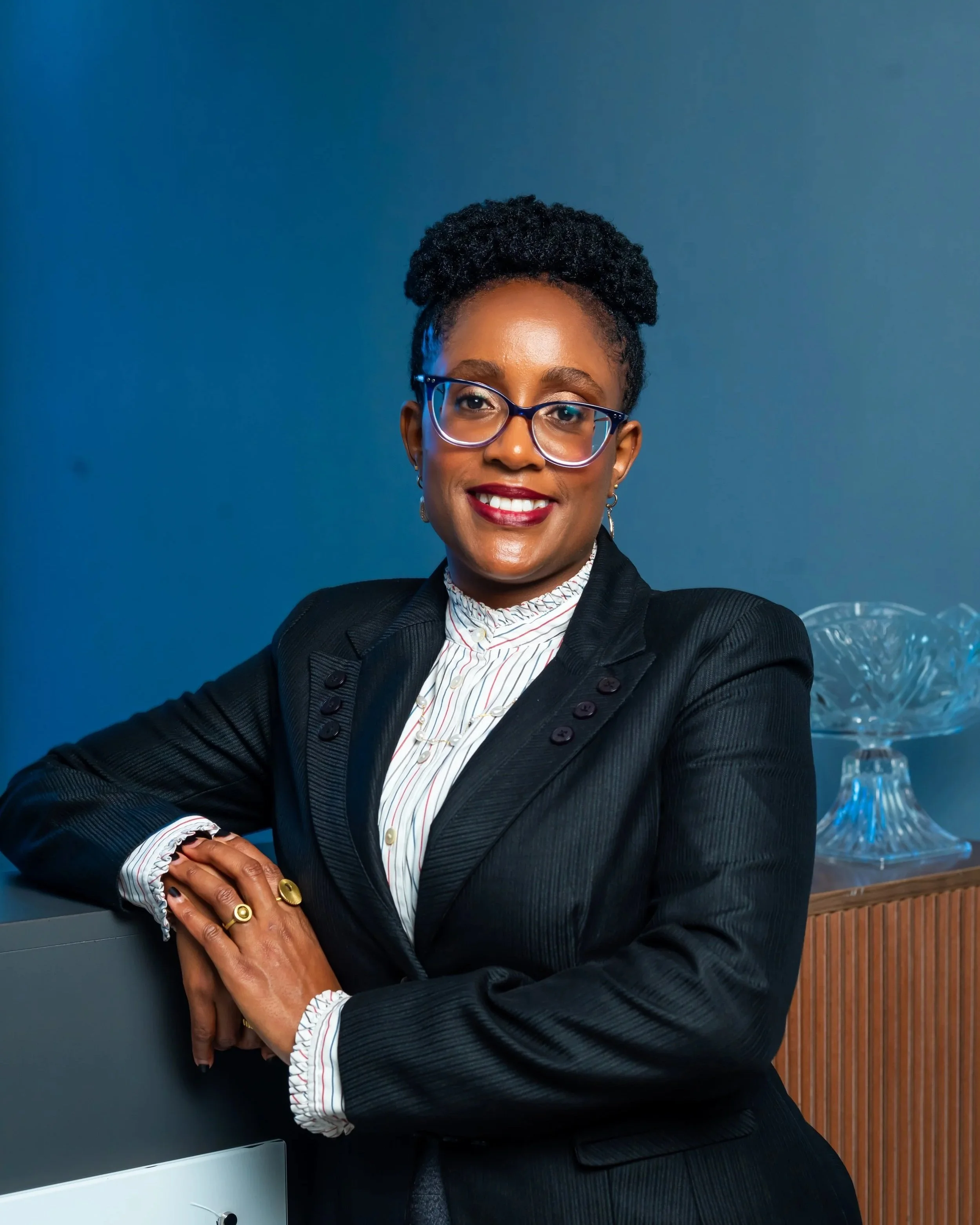 A professional woman with glasses and earrings, wearing a black blazer and striped blouse, smiling with arms crossed in an office setting with a blue wall and glass bowl in the background.