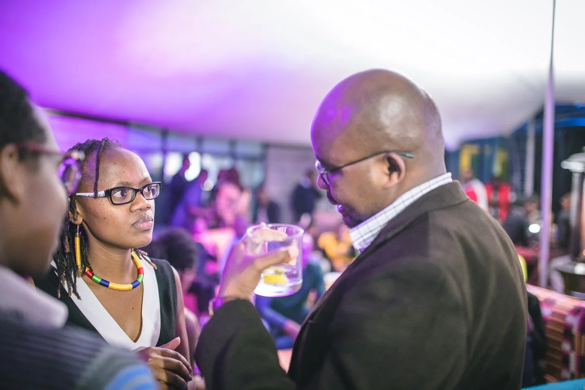 Three people engaged in conversation at a social event, with one holding a drink, under purple lighting with blurred background.