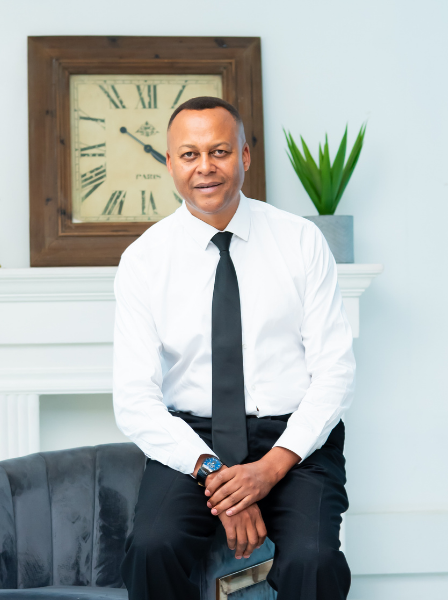 Kairo Thuo, Viva's Managing Partner in white shirt and black tie sitting on a gray sofa in a room with a white wall, a large wall clock, and a green potted plant.