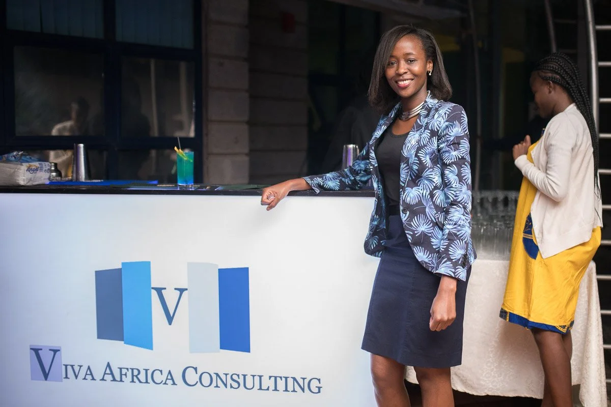 A smiling woman in a blue and white patterned blazer standing at a white counter with a logo for Viva Africa Consulting, holding the edge of the counter with her right hand.