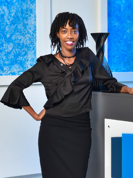A smiling woman in a black blouse and black skirt standing at a reception desk with a blue and white background and a black decorative vase.