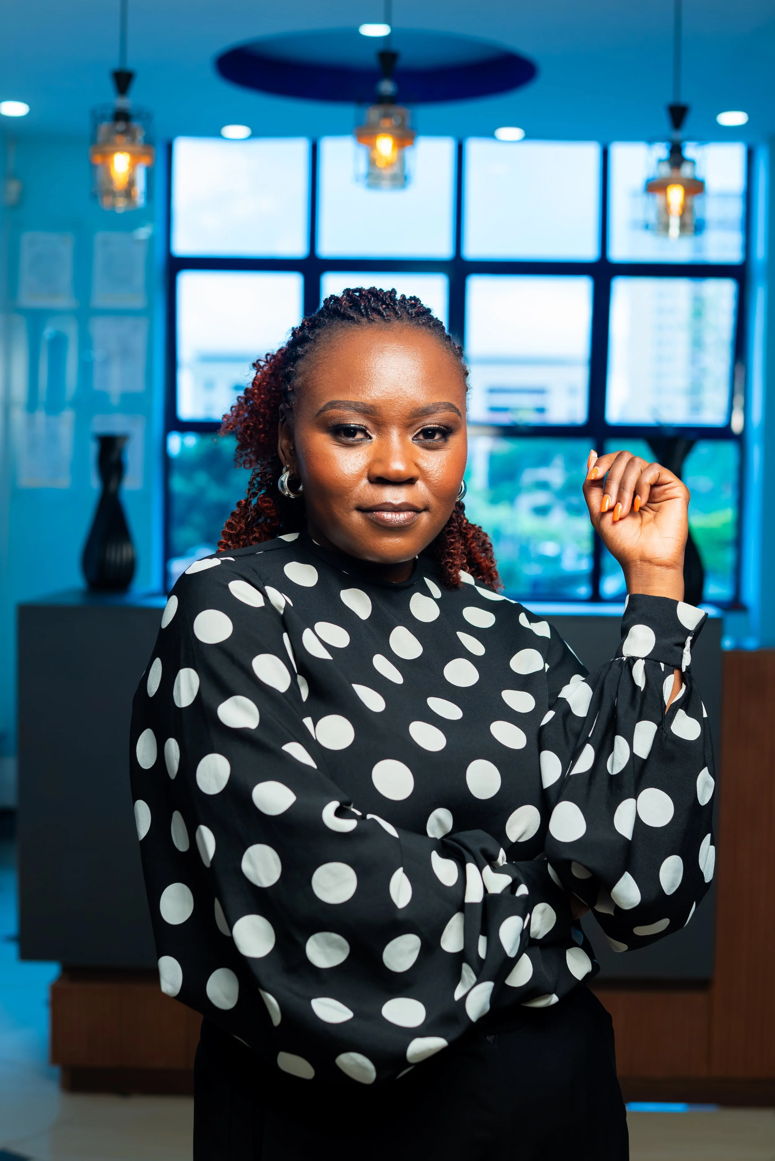 A confident woman with braids in a polka dot blouse, raising her fist in a modern office setting with large windows and pendant lights.