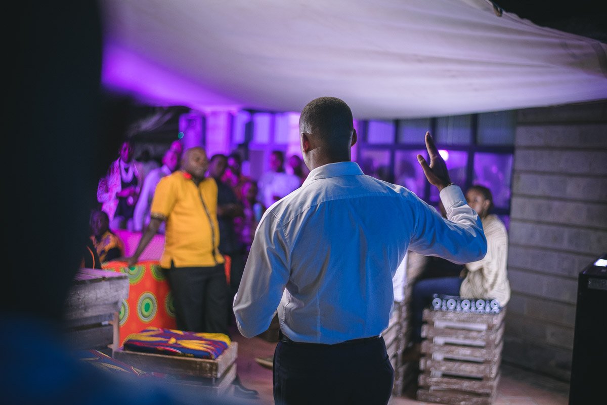 Kairo Thuo giving a speech or presentation at an indoor event with purple lighting, audience members are visible in the background.